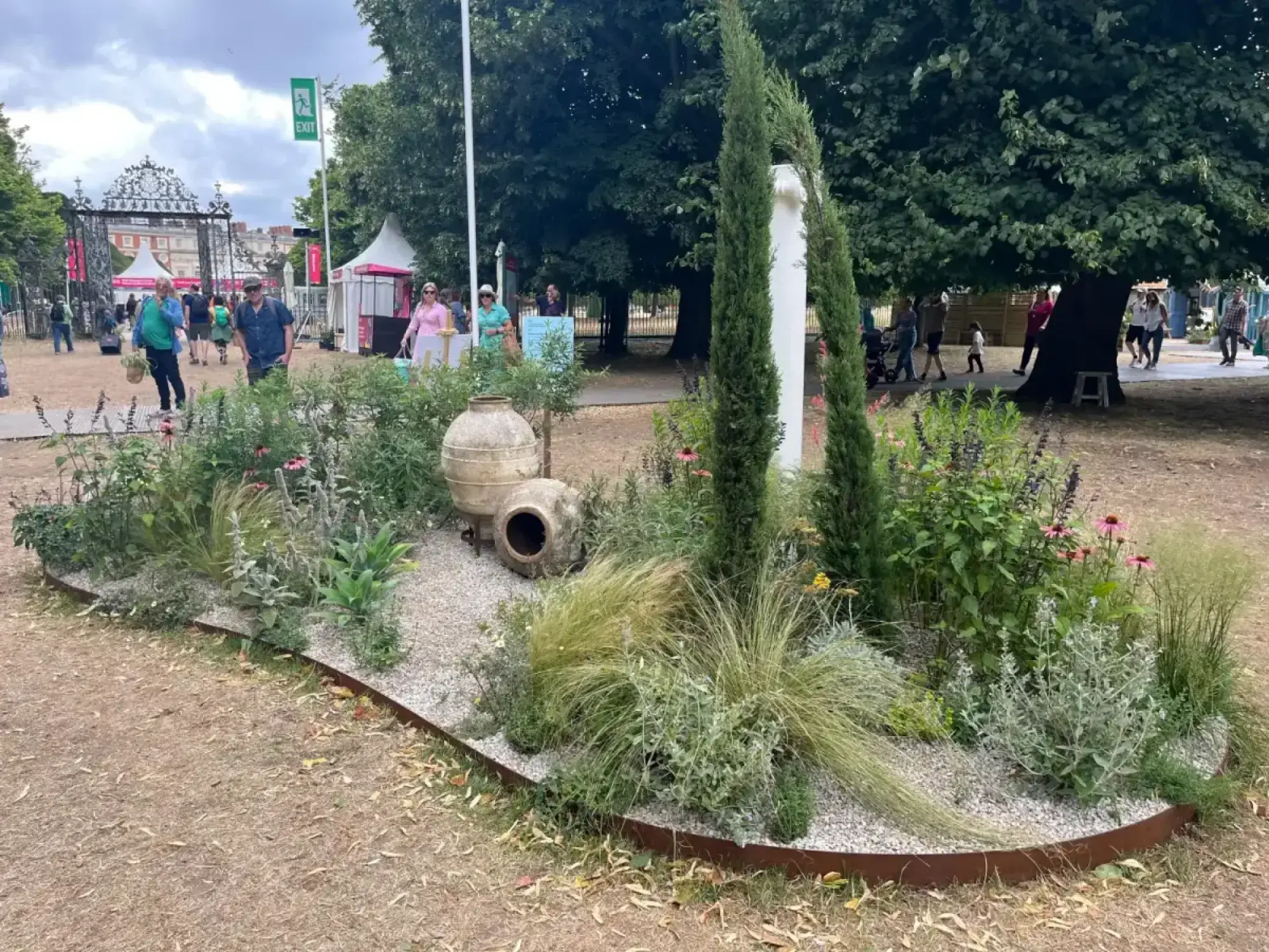 A feature border garden with two tall, slender conifers, a white abstract sculpture, and weathered terracotta amphorae, surrounded by ornamental grasses, pink coneflowers, and purple salvias on light grey gravel with rusted metal edging.