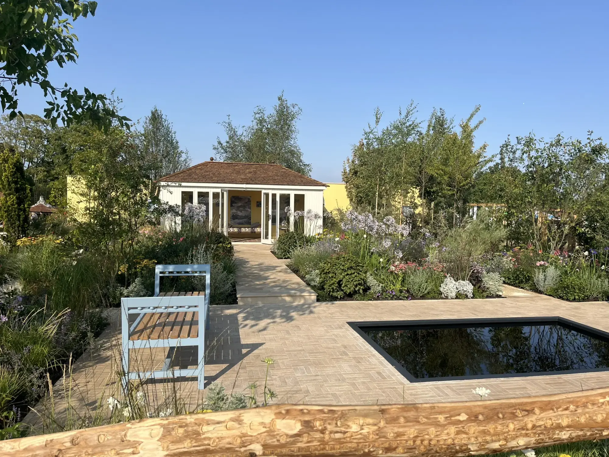 White pavilion, dark reflection pool, blue bench, and planted borders of agapanthus, grasses, and trees on light paving.