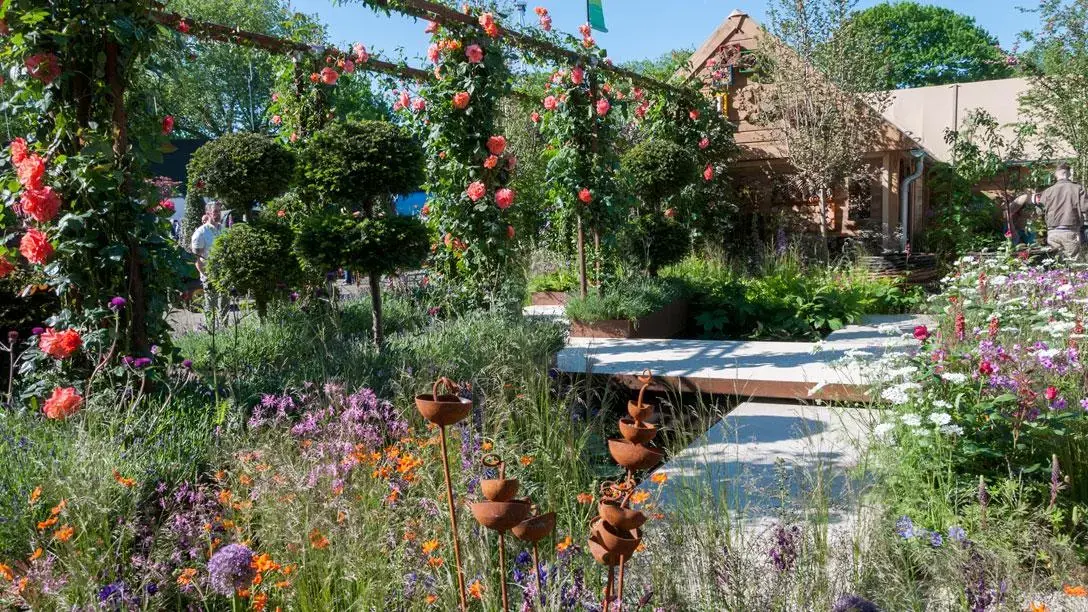 A vibrant feature garden with a coral rose-covered wooden pergola, naturalistic mixed planting, Corten steel water features, a concrete path, and a timber building.