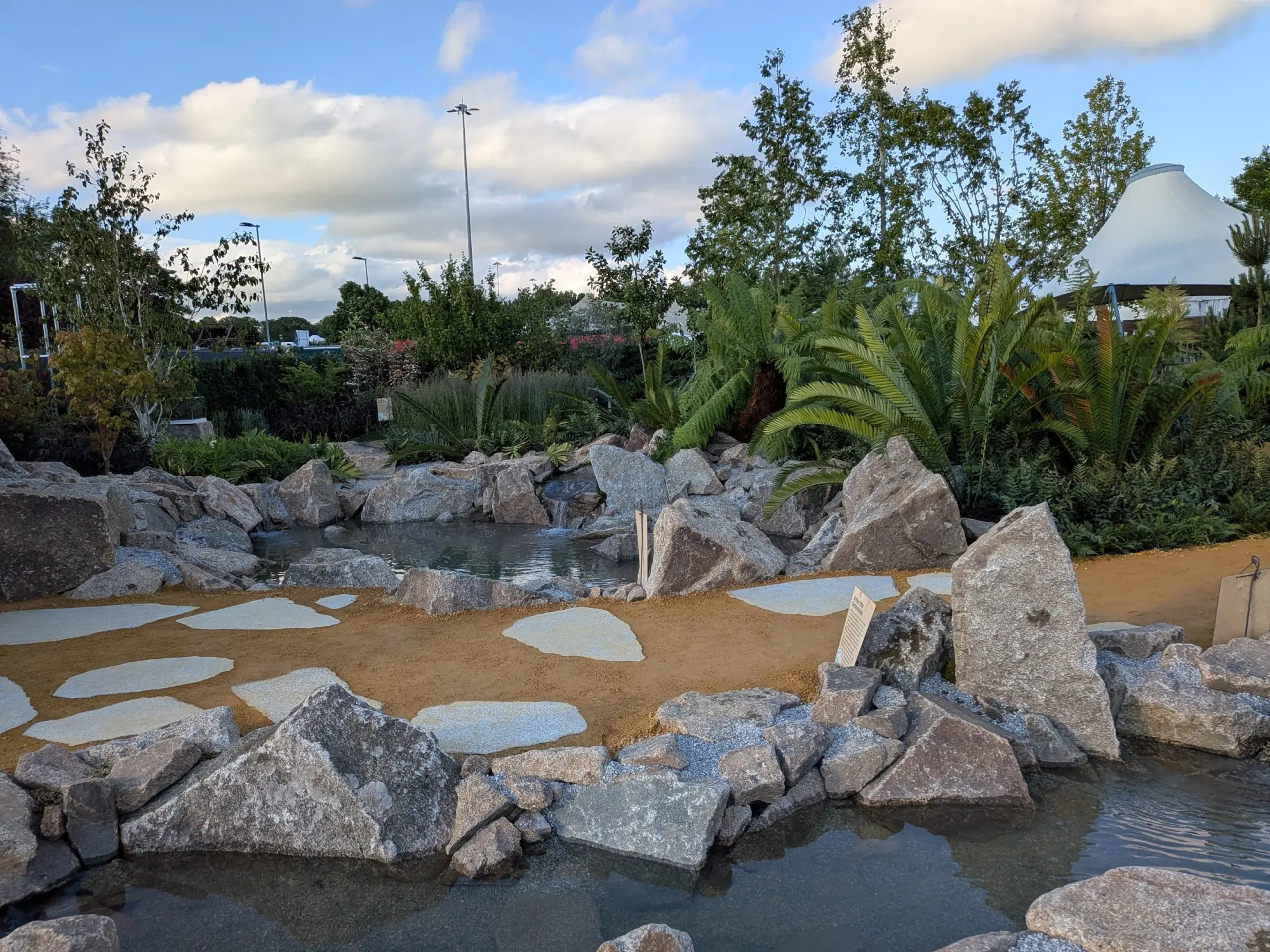 A naturalistic show garden features a winding stream with large grey boulders, light stepping stones on a sandy path, and lush green plants.