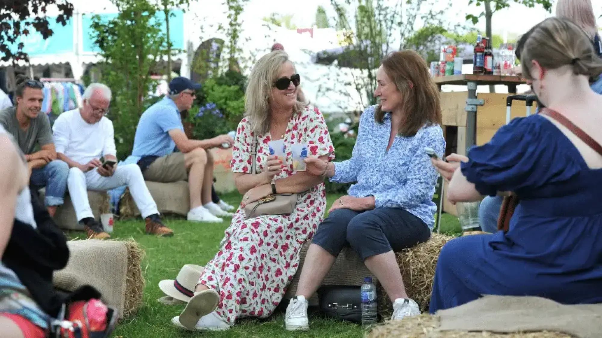 A casual outdoor garden featuring hay bales arranged for seating on a grass lawn, with a simple wooden bar structure in the background.