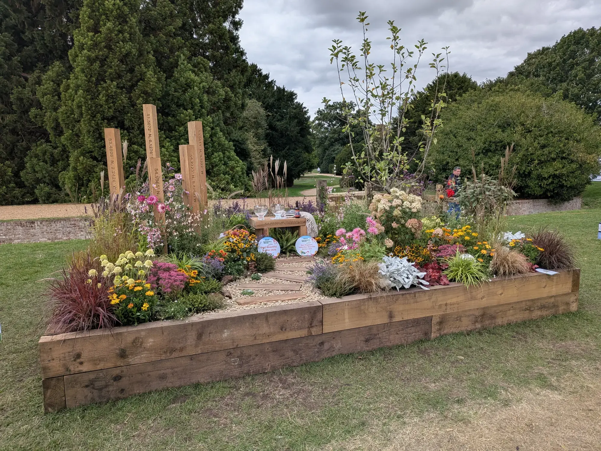 A wooden sleeper raised garden border contains a gravel path, a small wooden bench, colourful informal planting, and tall coordinate posts.