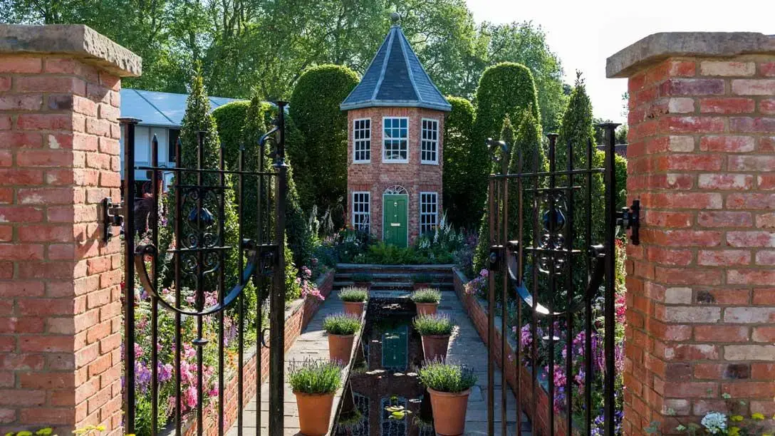 A whimsical show garden has a small brick tower with a green door, a central reflective rill, potted lavender, and colourful flower beds.