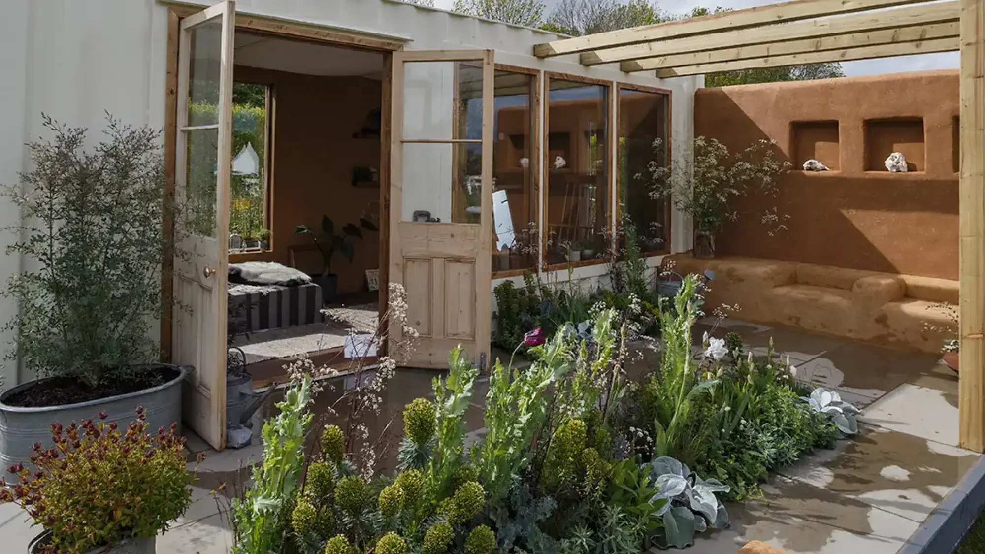 An artist's studio garden with a white building, open wooden doors, a warm earth-rendered wall with a built-in bench, and lush green planting.