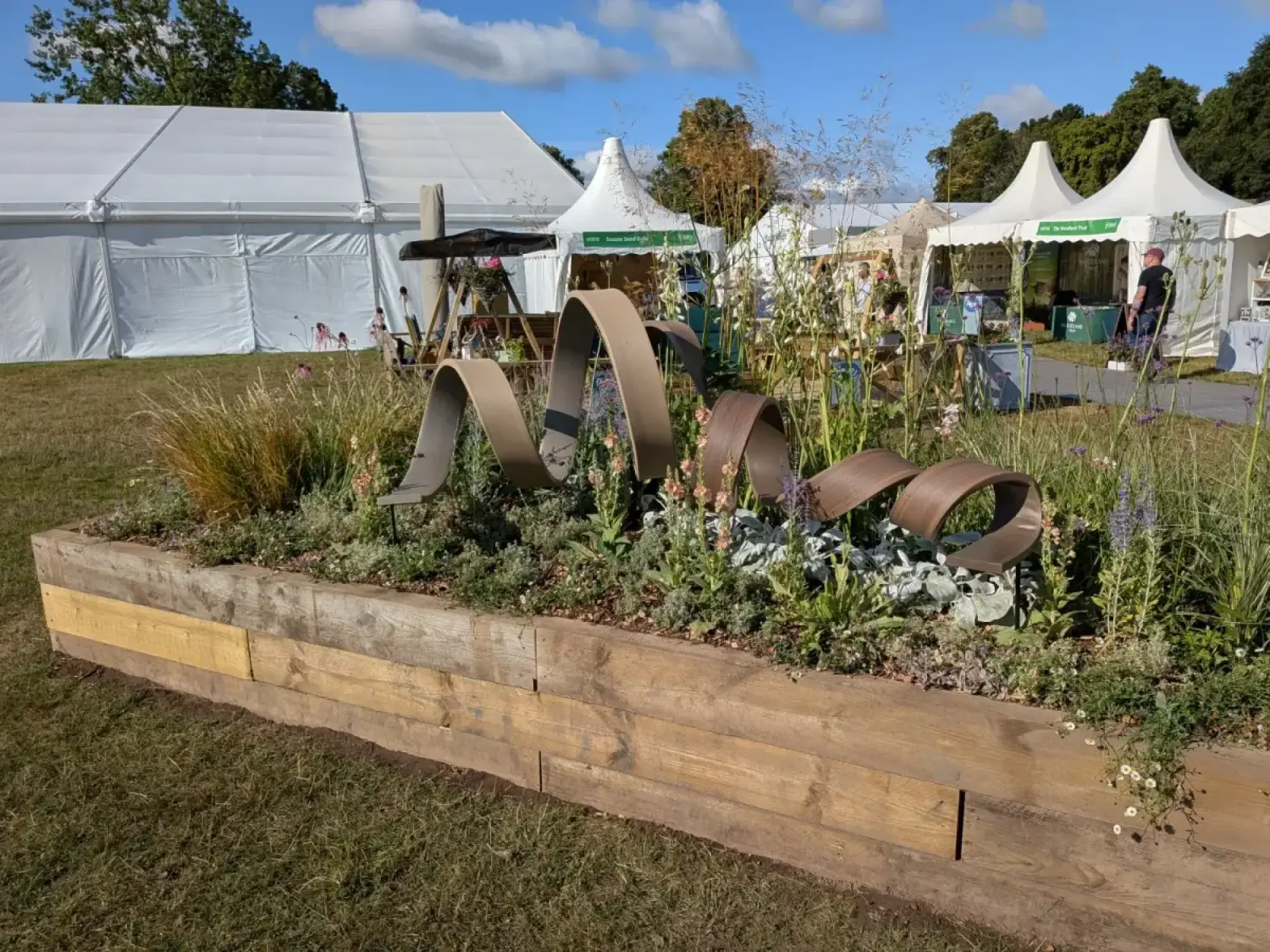 A raised timber long border garden features a wavy rusted metal sculpture among tall grasses, purple and peach flowers, and silvery foliage.