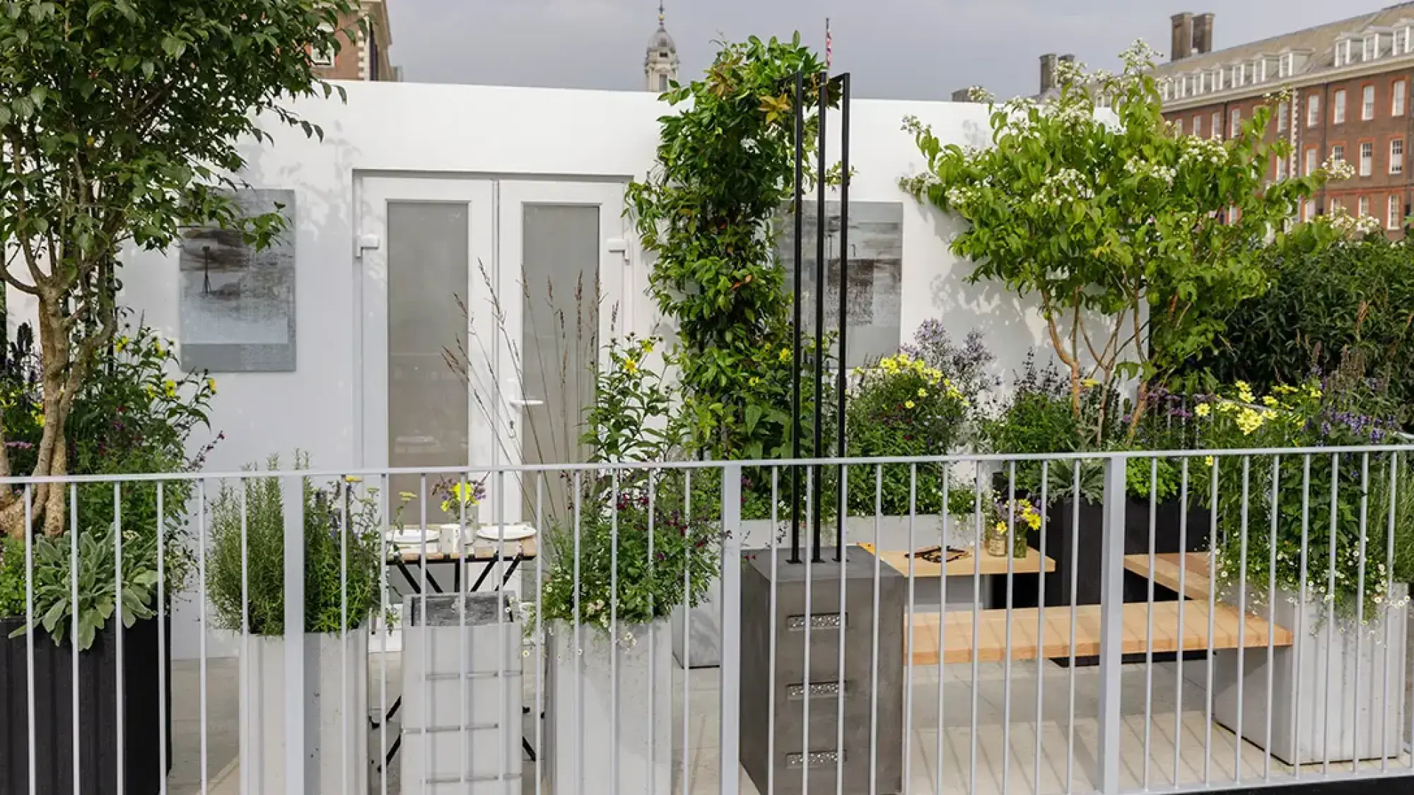 A modern balcony garden with large white and grey concrete planters, an integrated wooden bench, white double doors, and a black metal trellis with climbing plants.