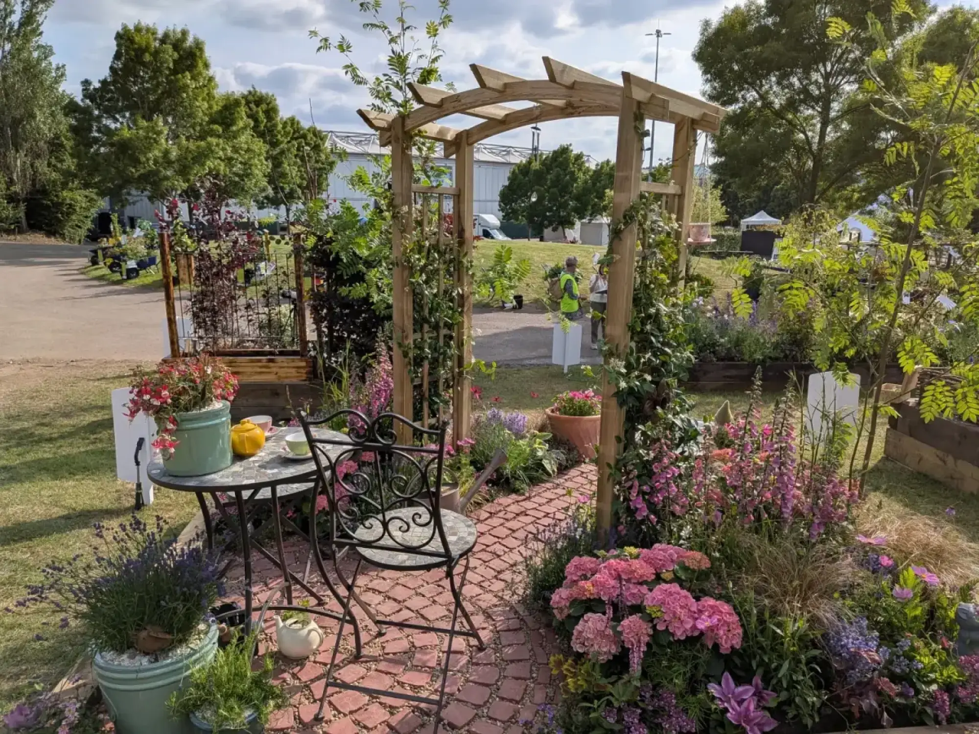 A charming border garden features a wooden pergola covered in climbers, a winding brick path, and a wrought iron bistro set surrounded by pink hydrangeas, purple foxgloves, and lavender.