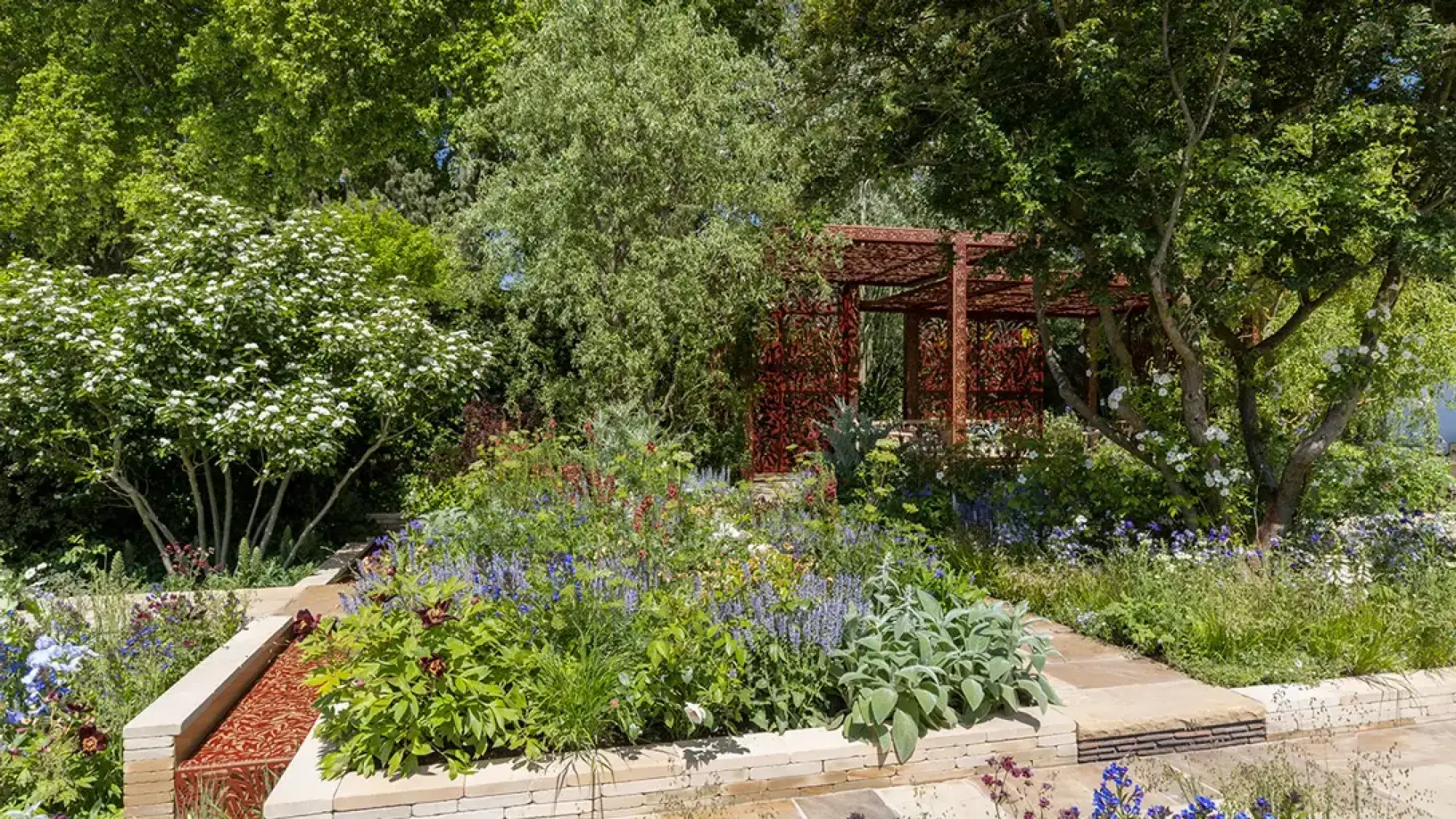 A lush show garden showcases an ornate red metal pergola, tiered stone walls with a patterned rill, and a vibrant mix of white, blue, purple, and red flowers.
