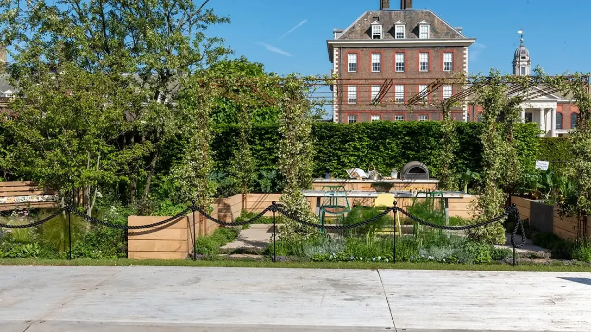 A sanctuary garden with wooden raised beds of edible plants, a pizza oven, and plant-covered archways defining an outdoor dining space.