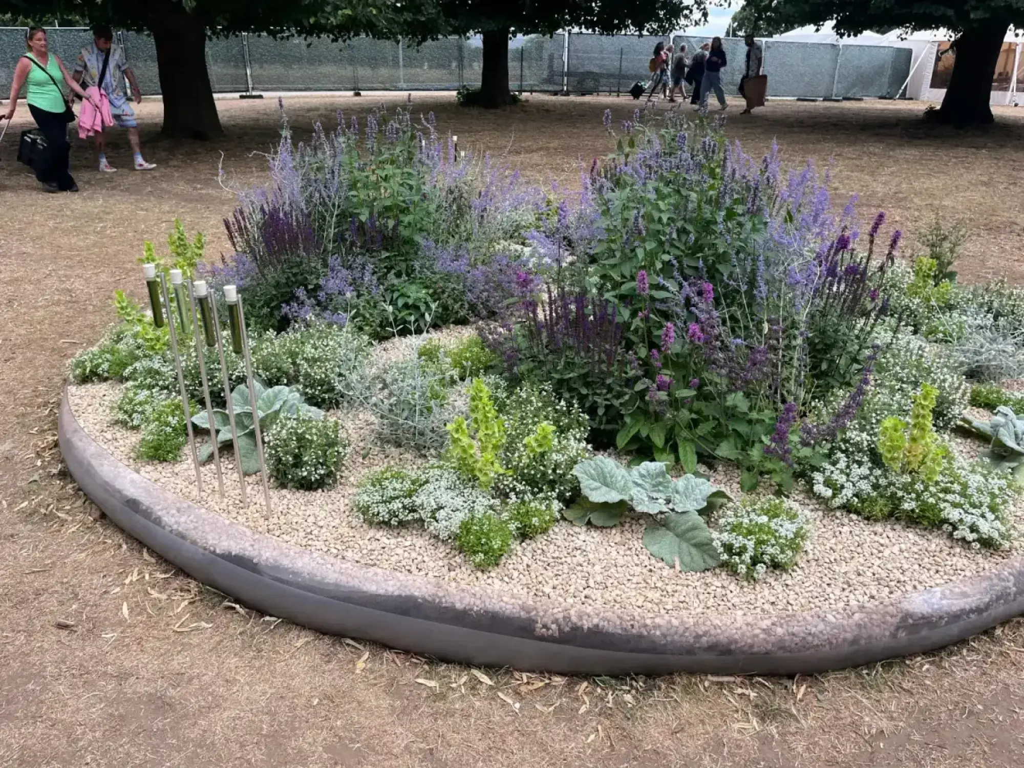 A curved, raised border garden filled with a variety of purple, white, and silver plants, accented by modern vertical tube sculptures and a gravel surface.