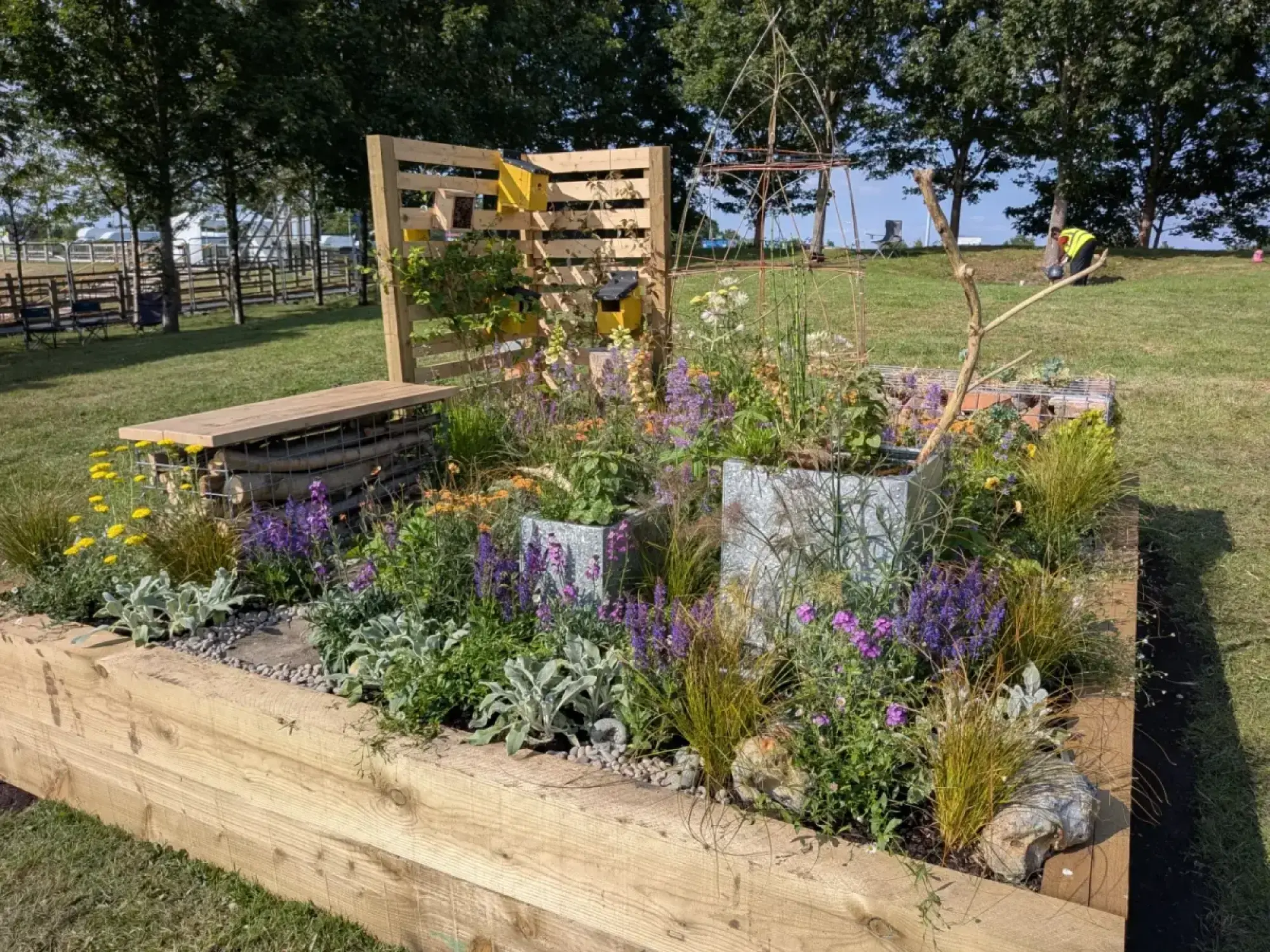 A beautiful border in a timber raised bed features a pallet screen with insect hotels, a gabion bench, and many colourful plants.