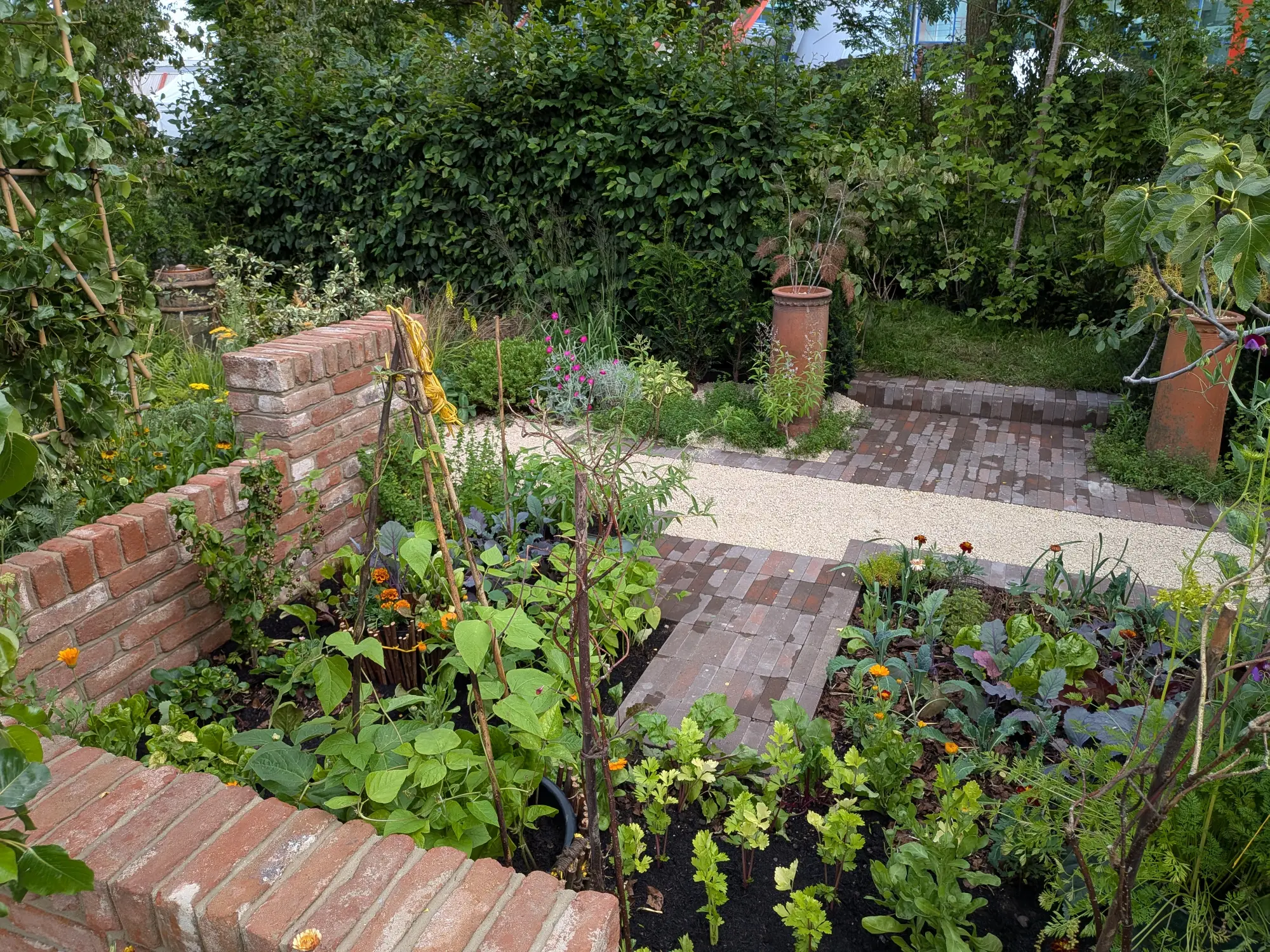 A productive kitchen garden features red brick raised beds and a gravel path, filled with leafy greens, orange calendula, and espaliered fruit.