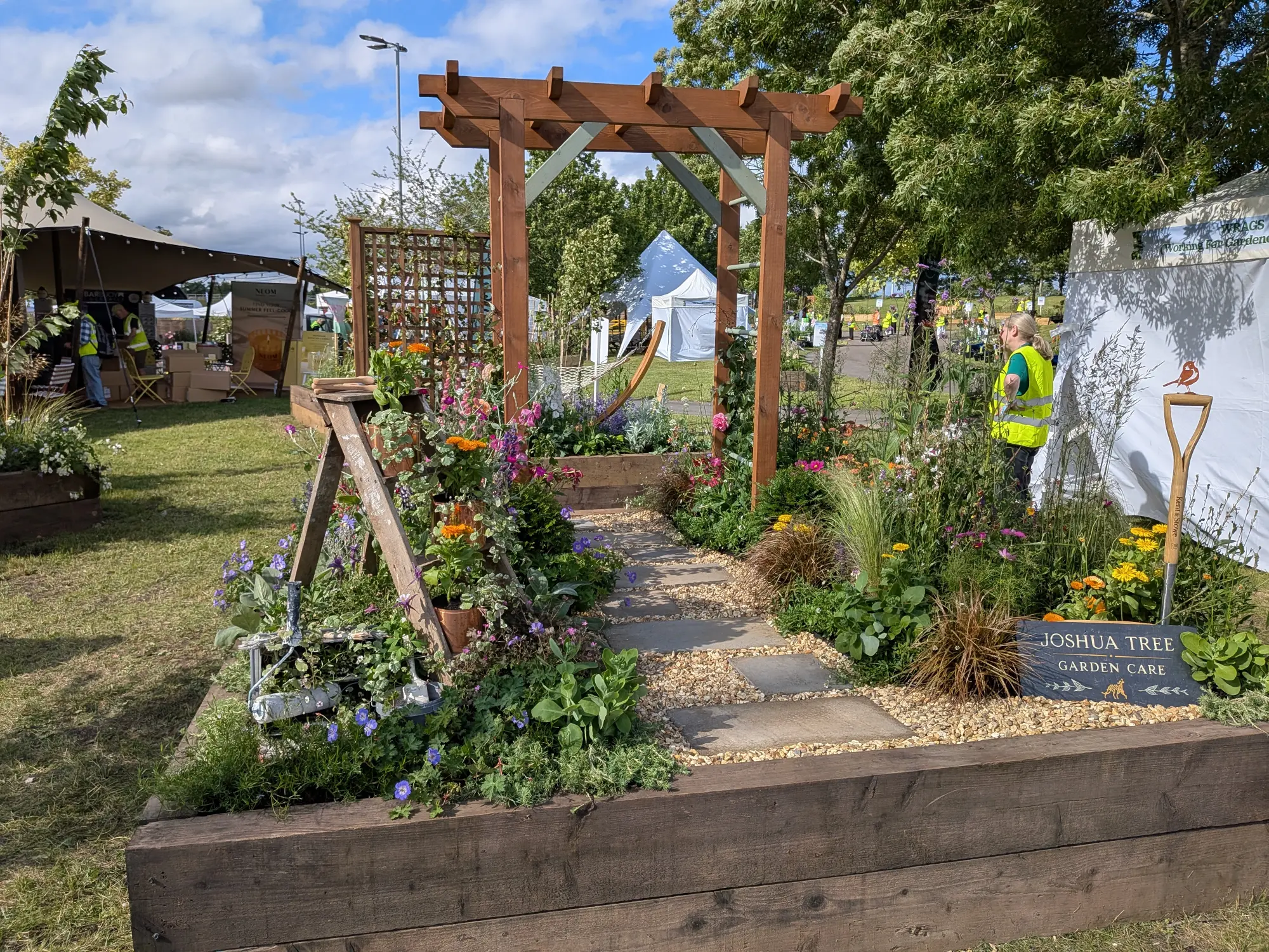 A creative garden border with a wooden pergola over a gravel path, featuring an artist's easel with a paint roller and a spade topped with a metal robin sculpture, surrounded by colourful mixed planting.