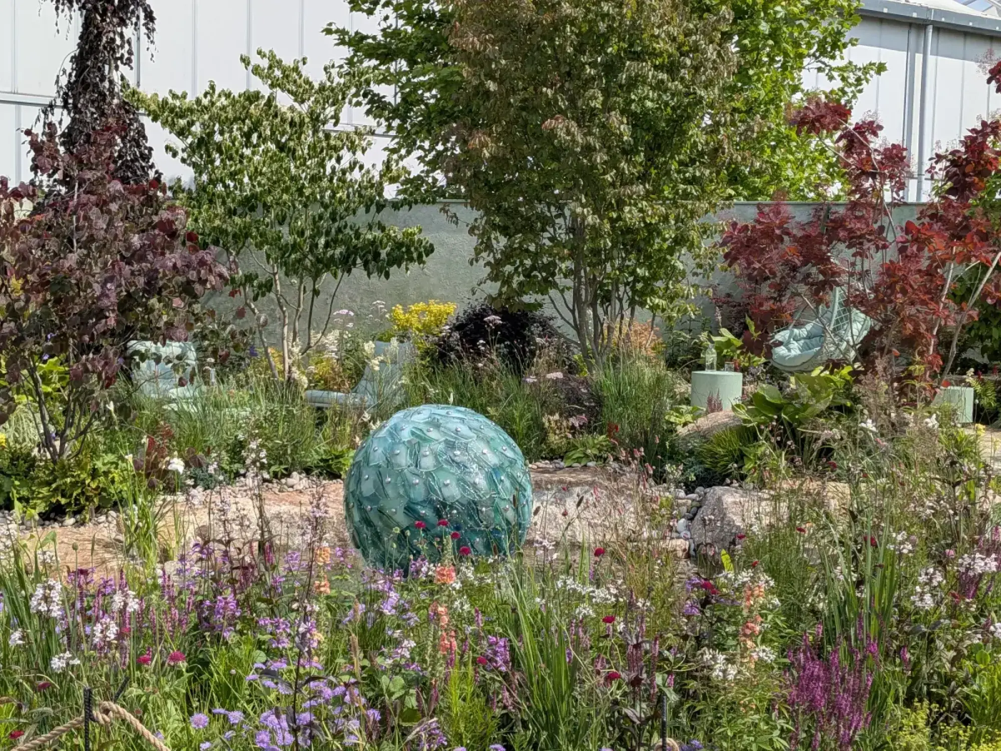 A show garden displays a large teal glass sphere sculpture amidst mixed perennial planting and trees, with aqua hanging chairs.