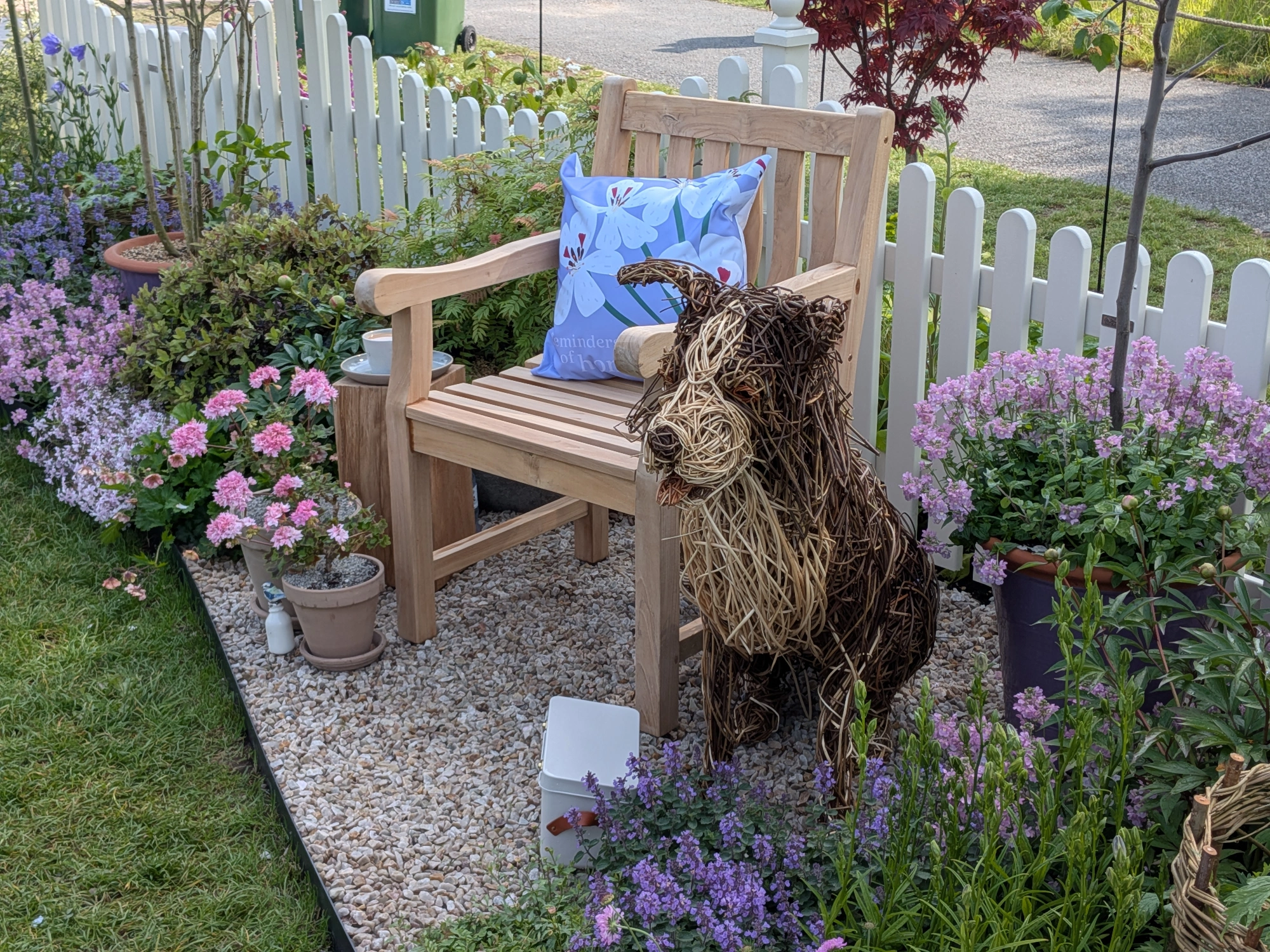 A charming cottage border displays a wooden bench, woven dog sculpture, and purple and pink flowers beside a gravel path and white picket fence.