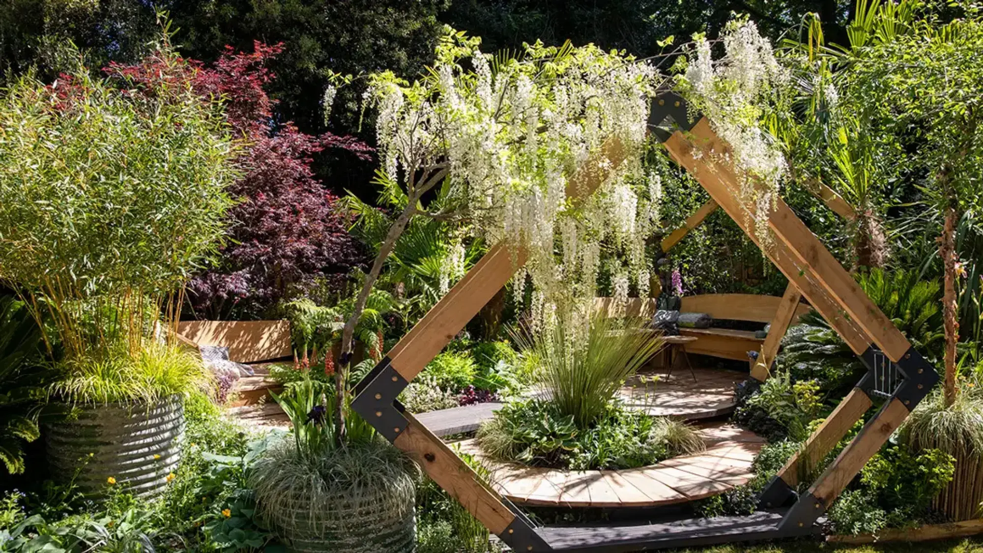 A serene sanctuary garden features a white wisteria-draped angular timber archway, framing a circular wooden deck with benches and lush green plants.