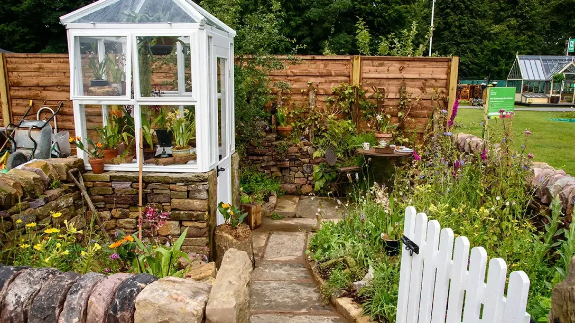 A charming terrace garden features a white greenhouse filled with orchids, a winding flagstone path, dry stone walls, mixed cottage garden planting, and a small white picket gate.