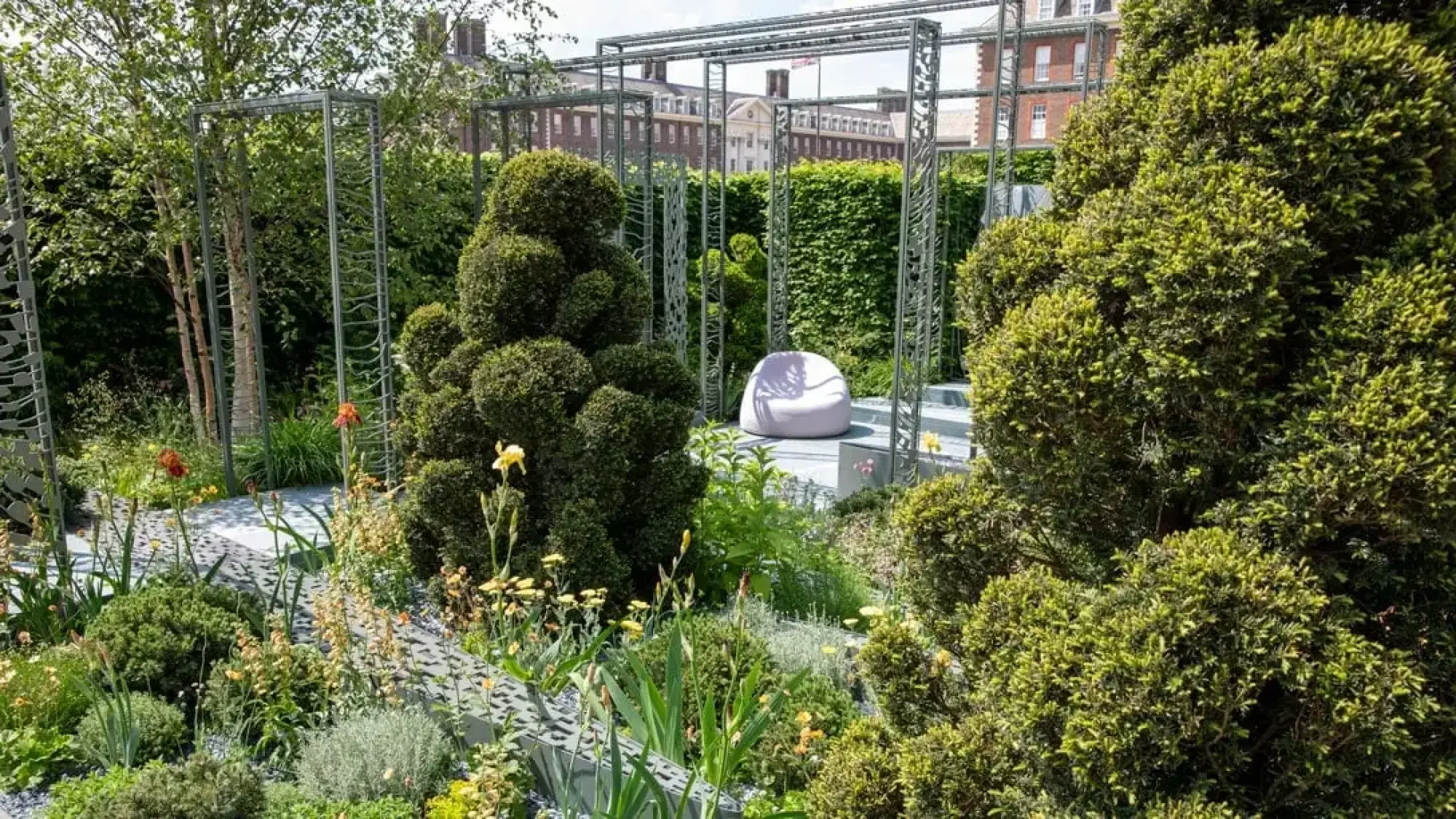 A contemporary sanctuary garden with laser-cut metal pergolas, rounded green topiary, a lilac beanbag chair, and a textured grey path.