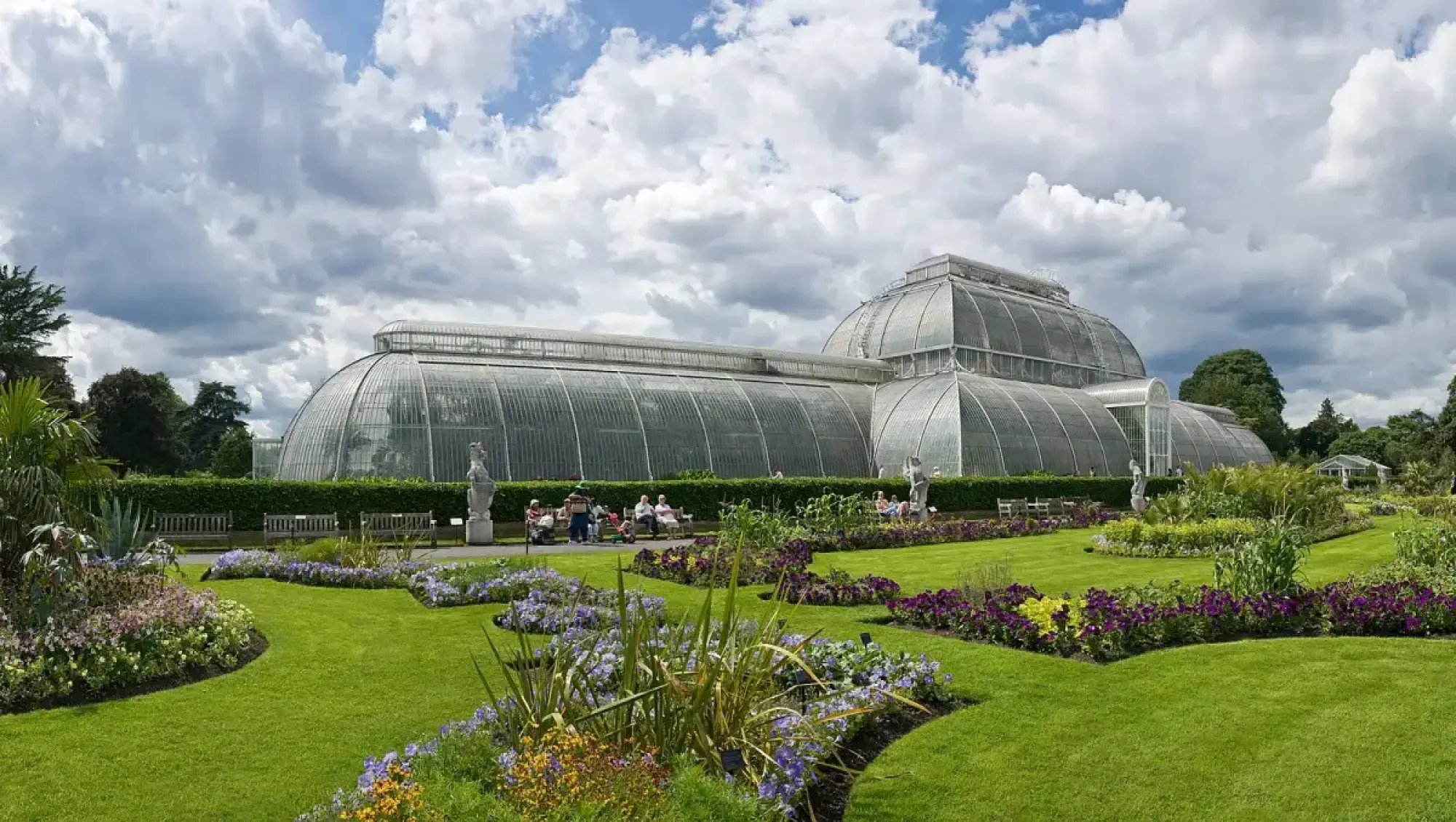 A vast, curved Victorian glasshouse with a white framework overlooks manicured green lawns and colourful flower beds filled with purple, blue, and yellow blooms.