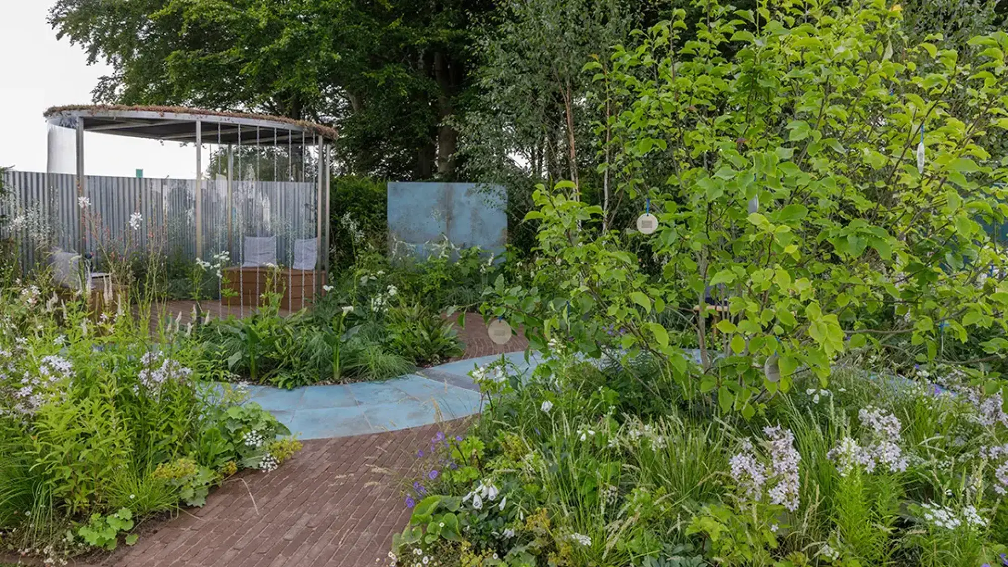 A contemporary garden features an open metal shelter, a light blue path, lush naturalistic planting, and a wooden bench.