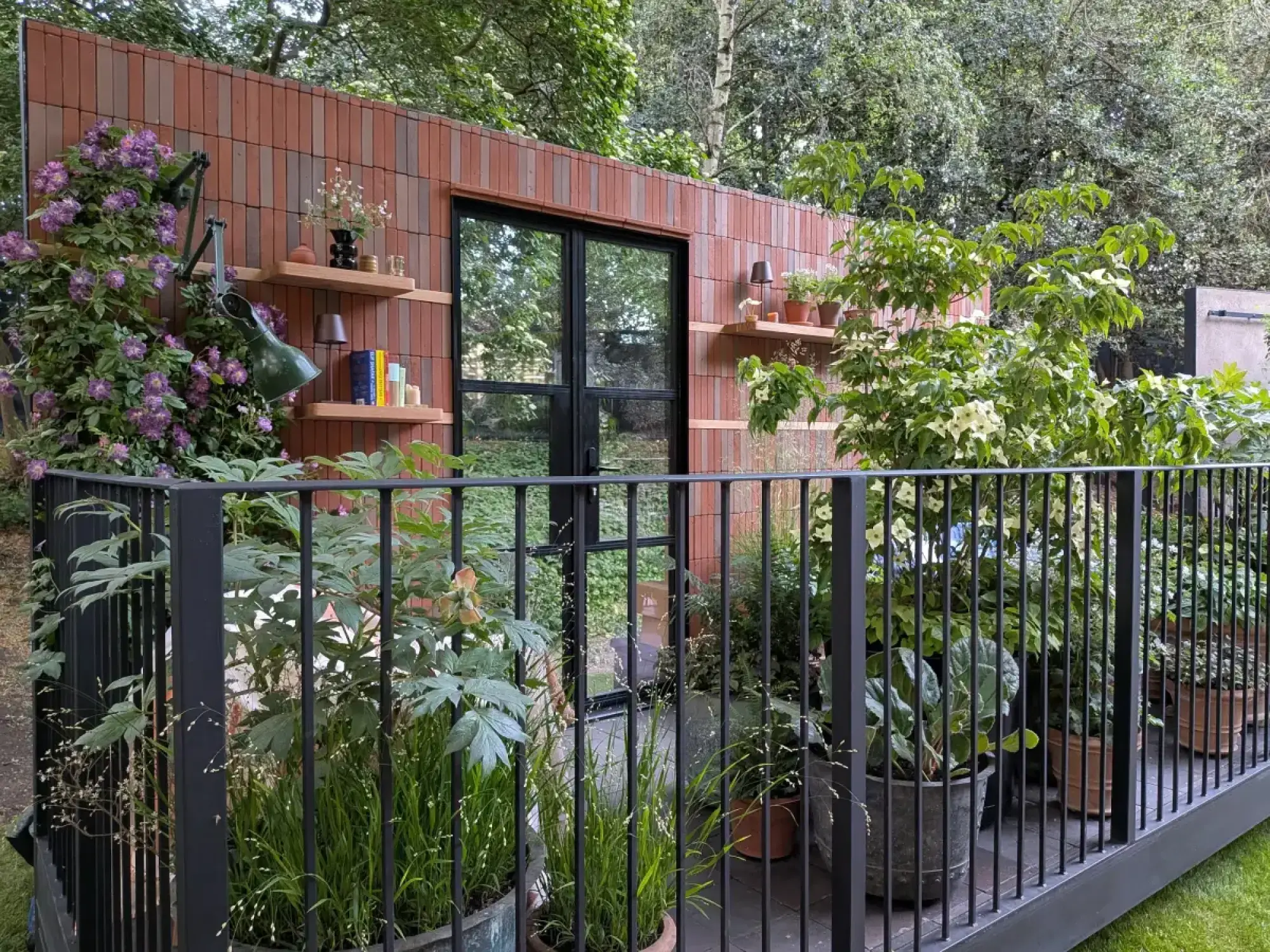 A contemporary balcony garden features a red brick wall with wooden shelves, books, a desk lamp, and black-framed doors, surrounded by potted plants.