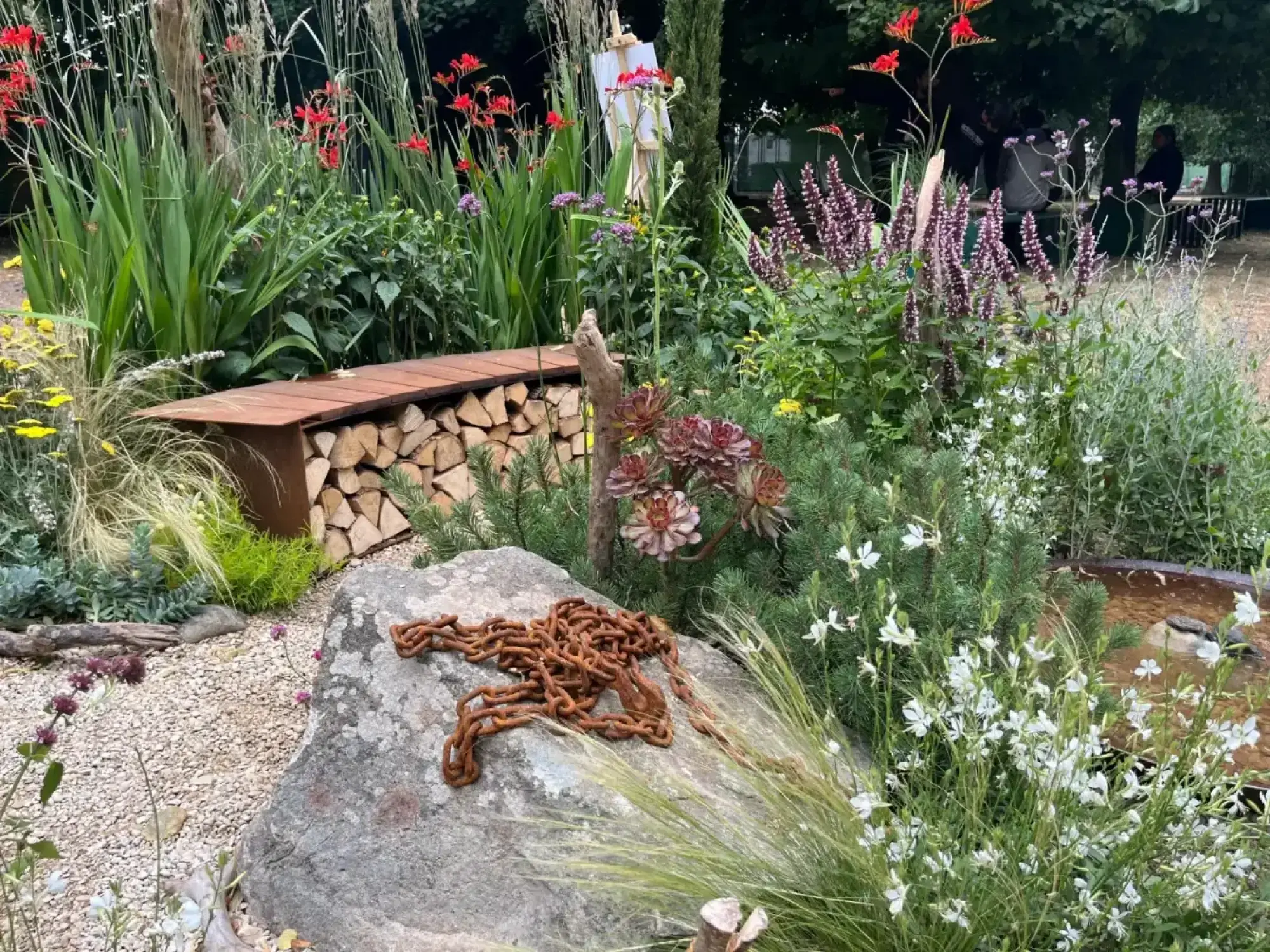 A naturalistic coastal border showcases a grey boulder with a rusted chain, red and purple flowers, white Gaura, grasses, and a corten steel wood store.