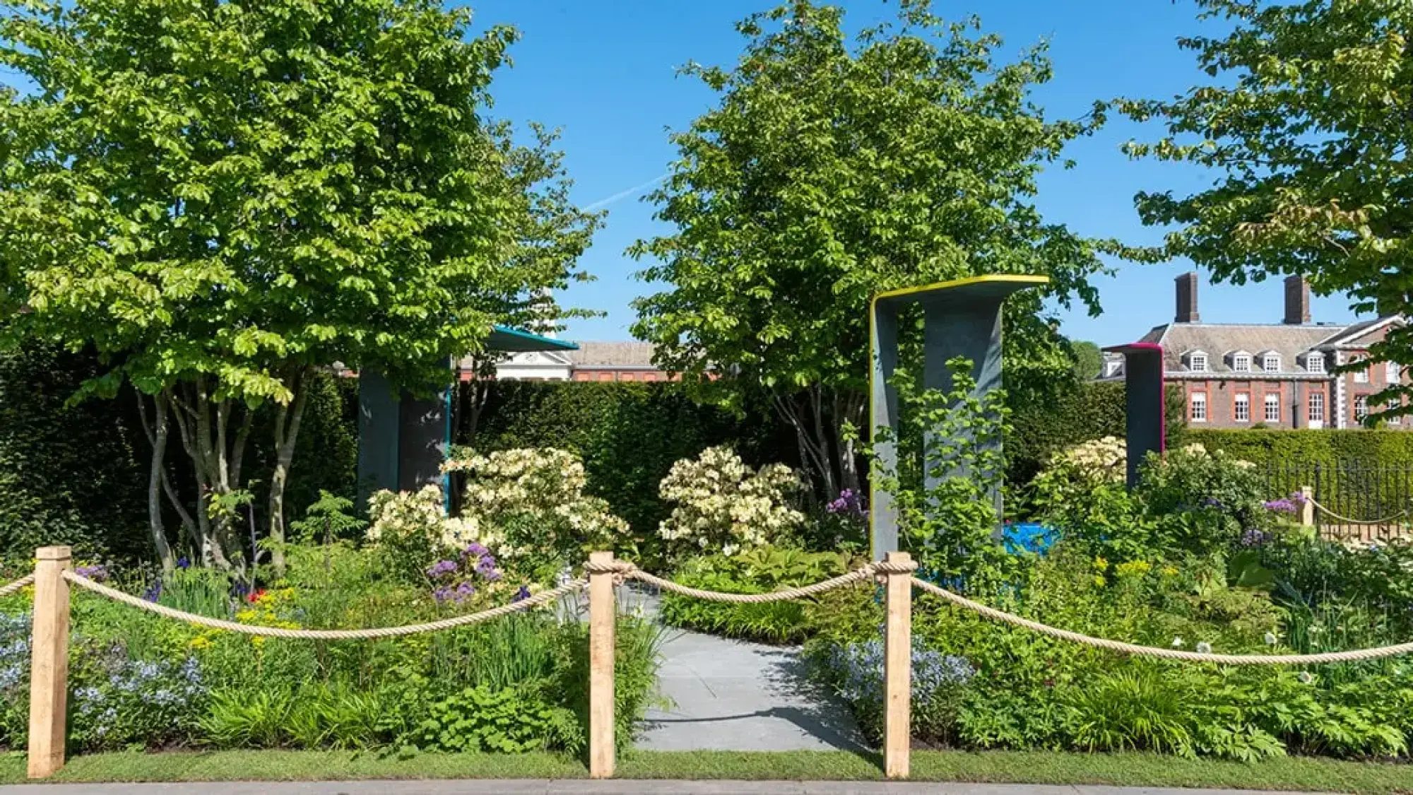 A contemporary sanctuary garden features two abstract, dark grey sculptural structures with bright yellow and pink caps, set among lush, naturalistic planting with a winding grey path.