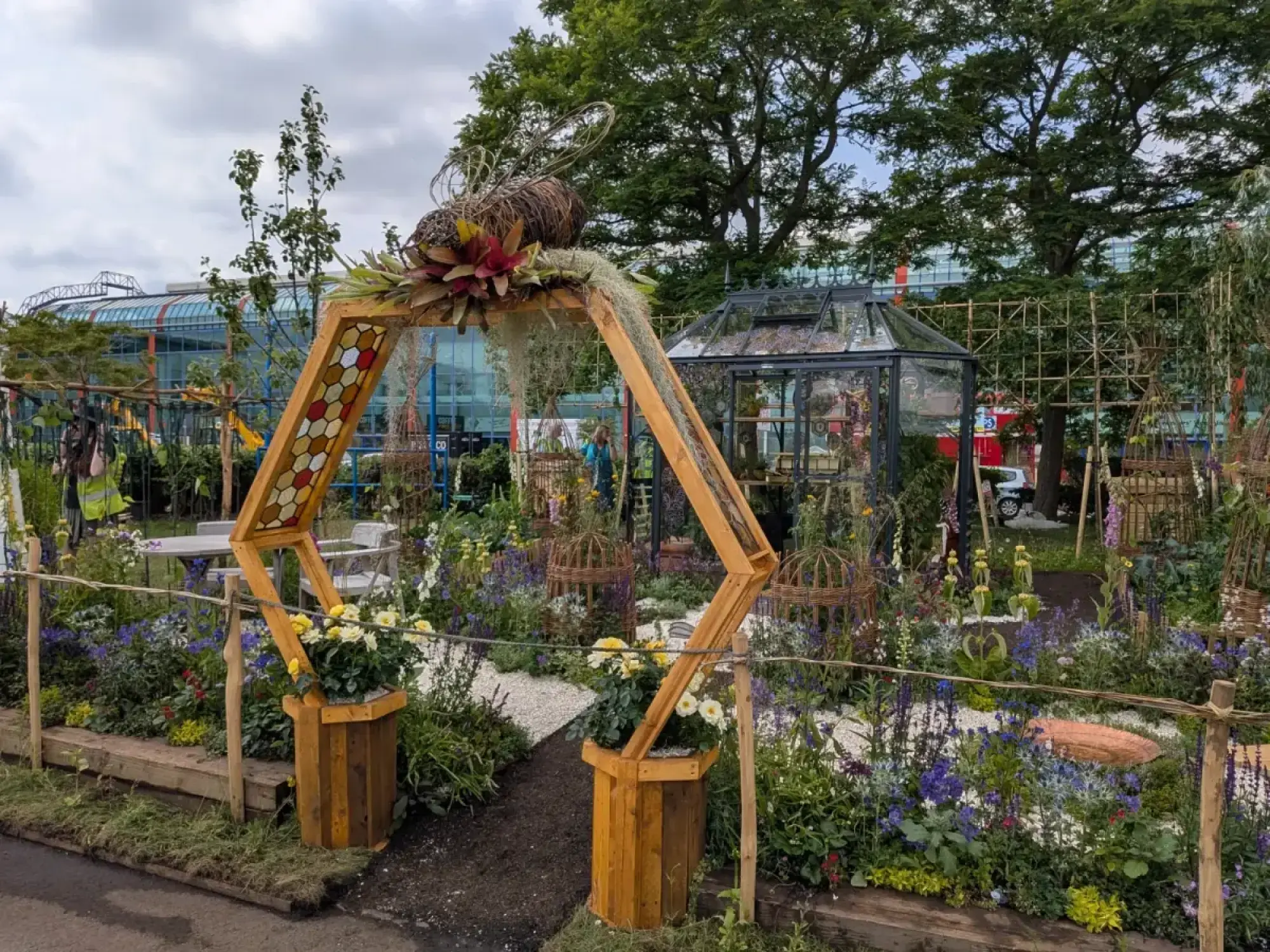 The Nectary showcase garden features a hexagonal wooden archway topped with a giant woven bee sculpture, leading to a vibrant bed of purple, blue, and yellow flowers with a small black greenhouse in the background.