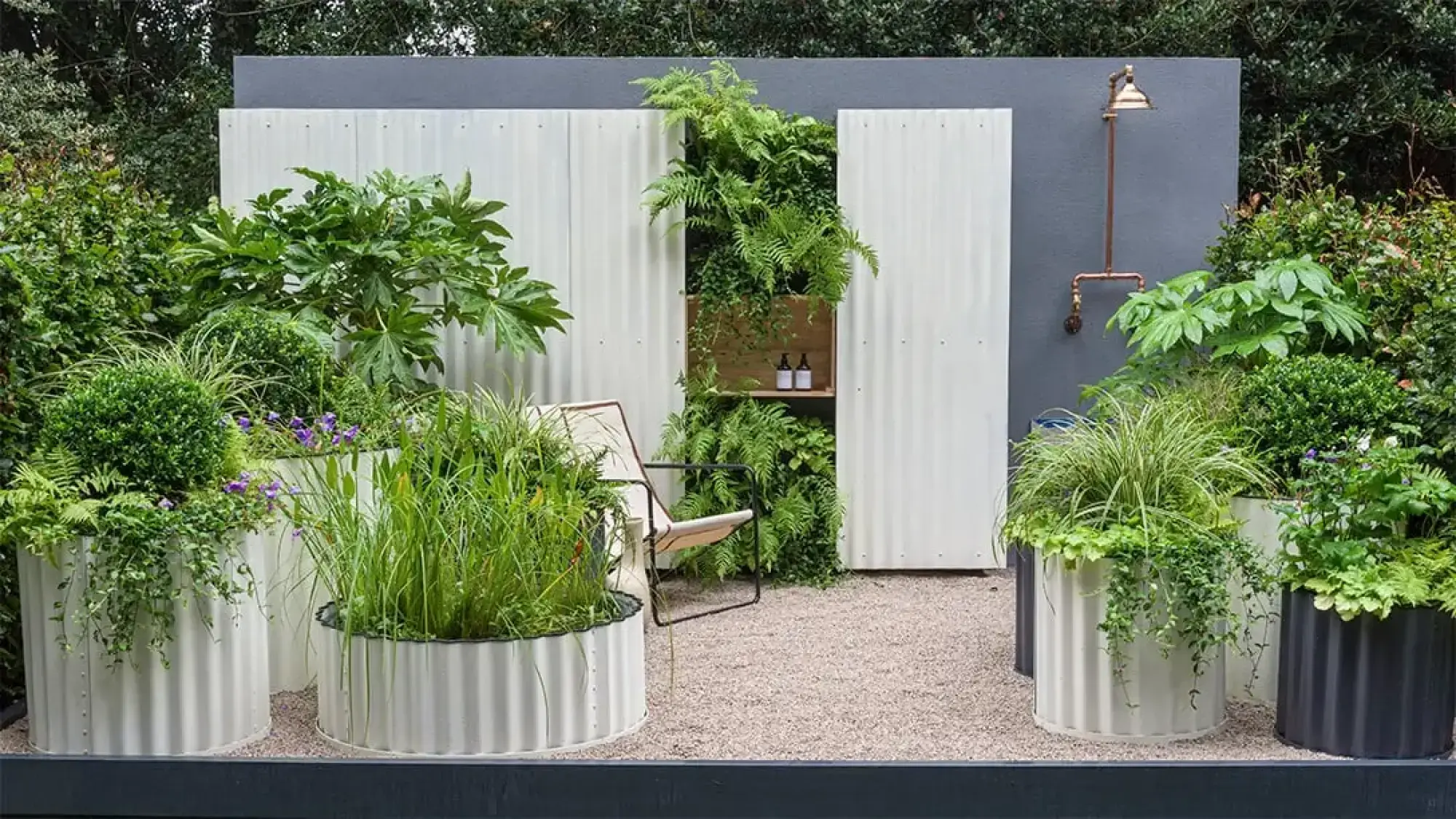A modern container garden with white and grey corrugated metal planters, a lush vertical fern wall, a timber-shelf niche, a simple lounge chair, and an outdoor copper shower.