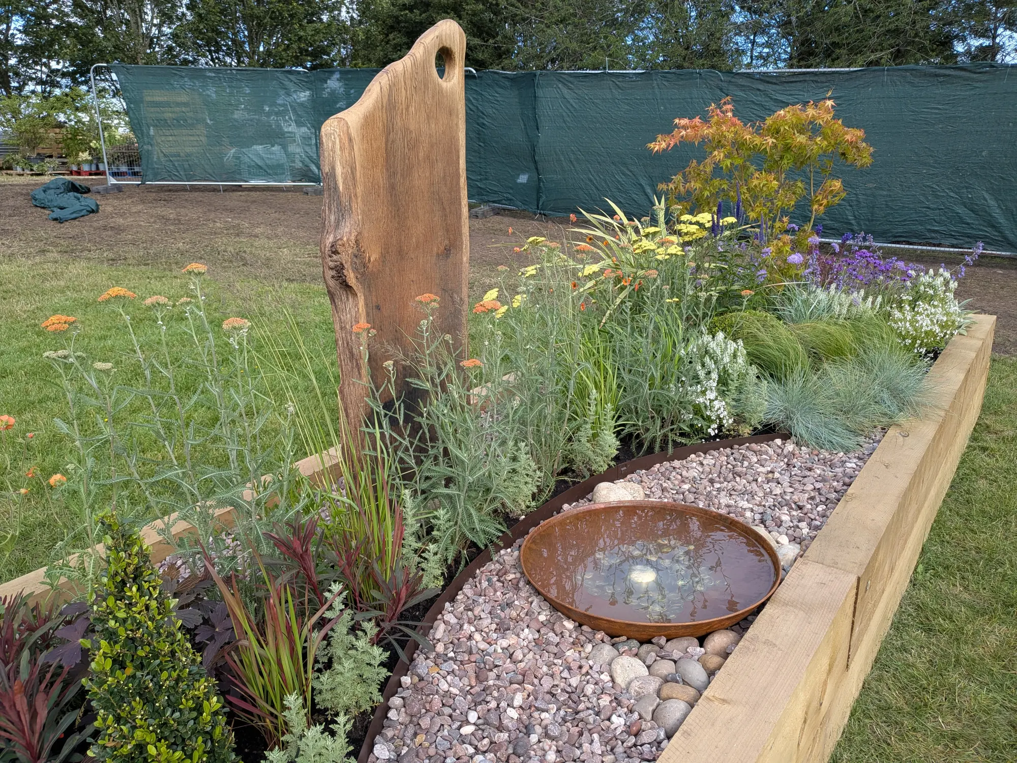 A contemporary garden border with a carved wood sculpture, a rusty water bowl set in gravel, and colourful naturalistic planting.