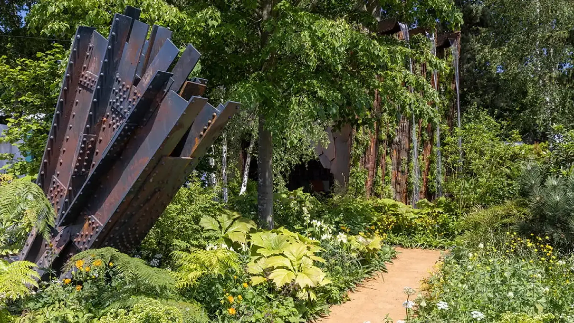A contemporary show garden displays a large, riveted, rusted steel beam sculpture among lush foliage, with a winding path and a vertical timber waterfall.