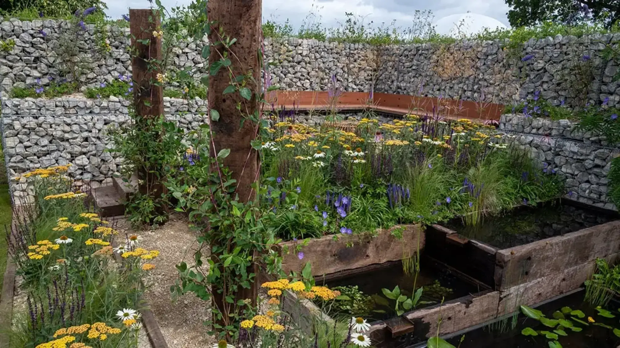 A city garden with stone gabion walls, tiered timber ponds, a curved bench, and a gravel path through wildflowers.