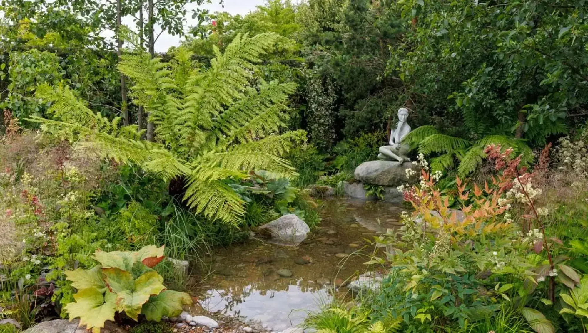 A serene woodland garden scene features a white female sculpture by a natural pond, amidst lush green tree ferns and diverse flowering plants.