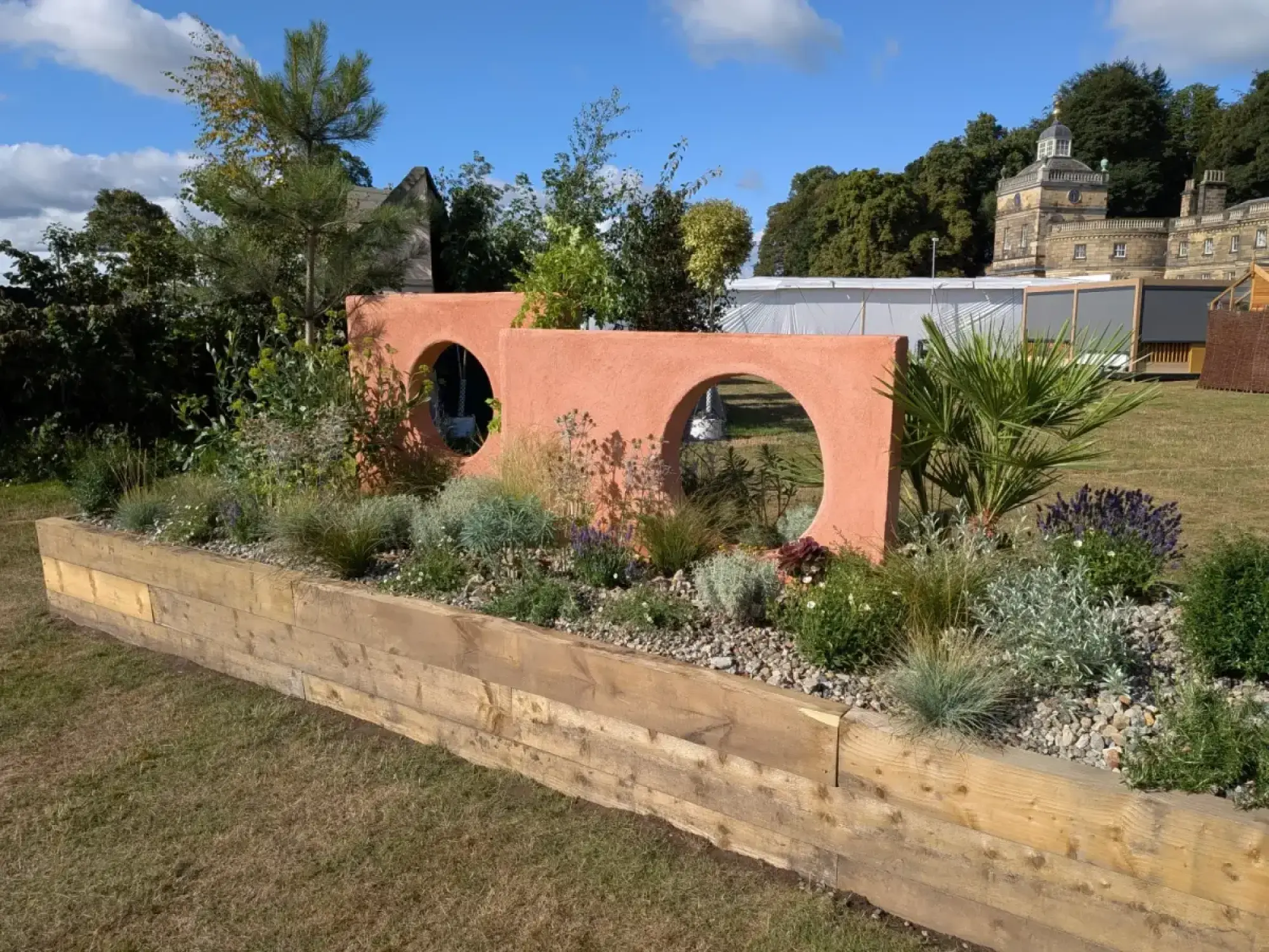 A long raised border garden features a terracotta-coloured wall with round openings, filled with drought-tolerant grasses, silvery foliage plants, and lavender, mulched with gravel.