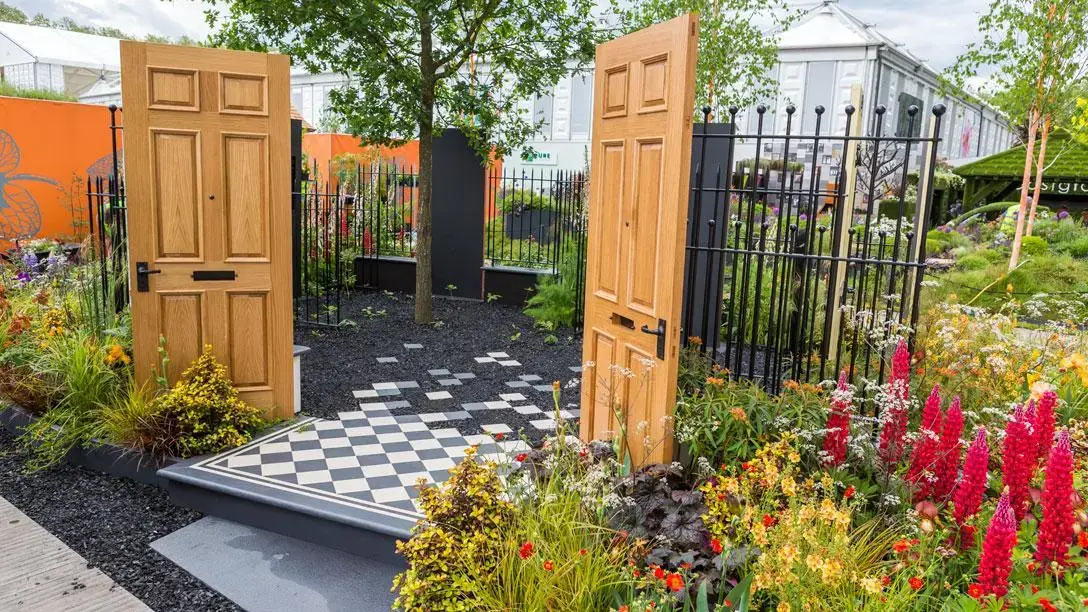 A conceptual Fresh Garden featuring two freestanding wooden doors and a black wrought iron fence, a checkerboard tiled path leading to a dark gravel area with a tree, and vibrant mixed planting with prominent red lupins.