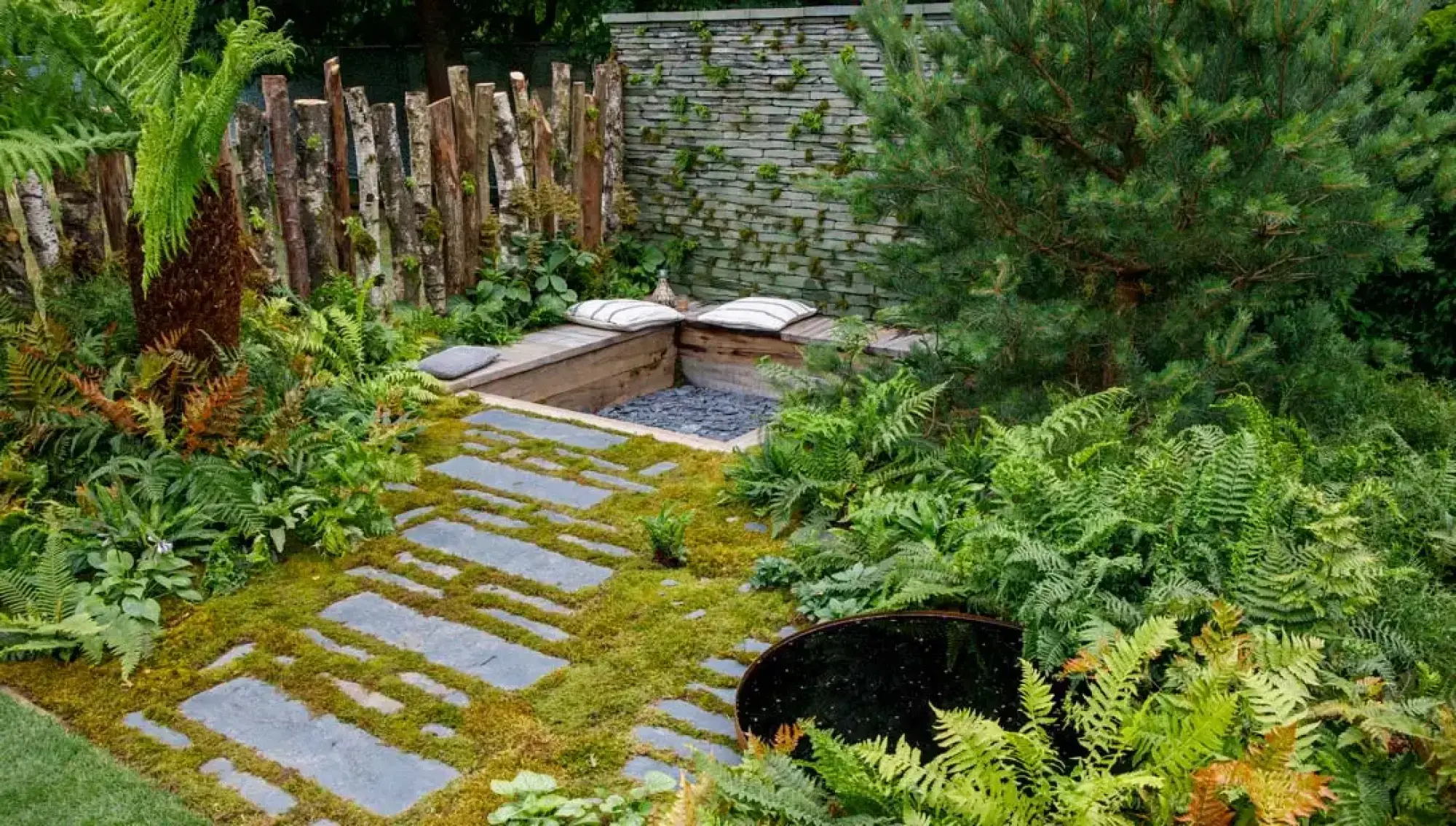 A tranquil naturalistic garden featuring a mossy path with stepping stones, a sunken wooden seating area with a gravel floor, a dark circular water feature, and various ferns under a pine tree.