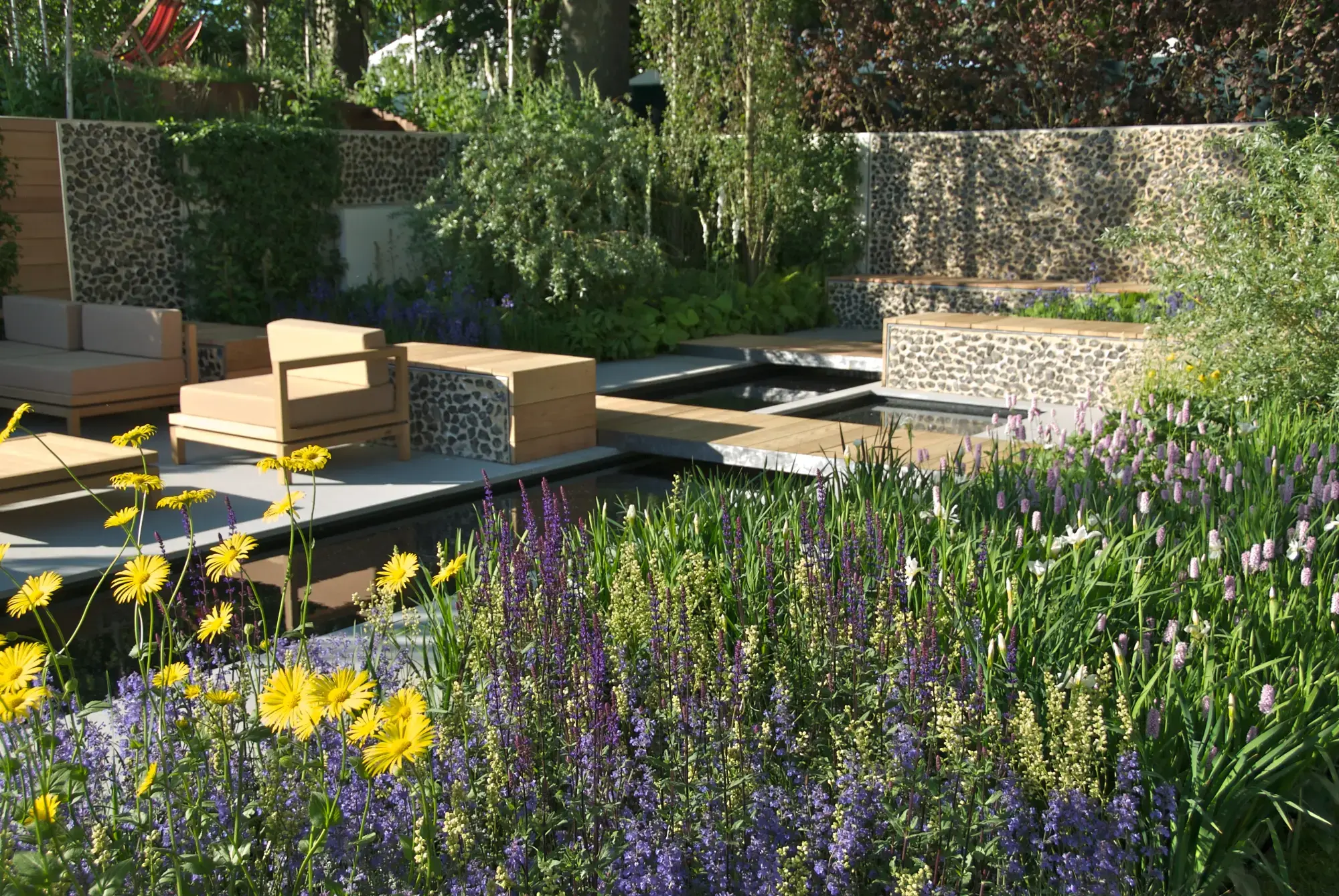 A contemporary show garden features linear water channels crossed by timber decking, surrounded by pebble-mosaic walls, modern wooden lounge seating, and vibrant purple, yellow, and pink flower beds.