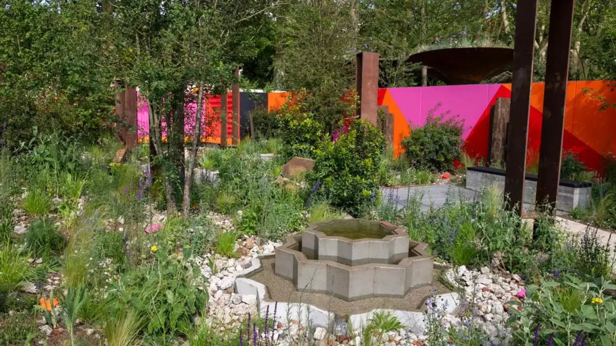 A contemporary feature garden with an octagonal tiered concrete fountain, vibrant geometric mural wall, rusted steel columns, and naturalistic planting.