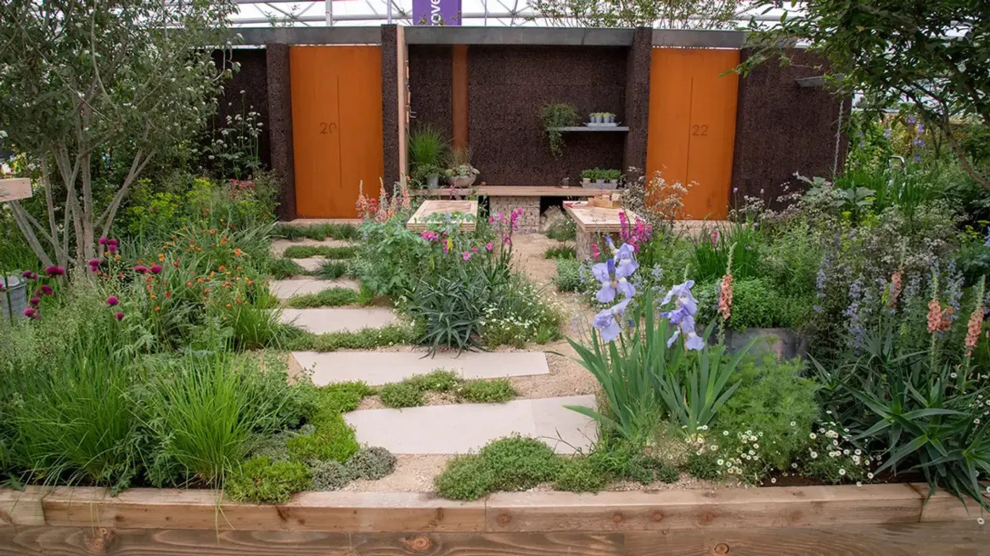 A naturalistic front garden with a stepping stone path through vibrant perennial planting, leading to a cork wall featuring rust-orange doors and gabion benches.