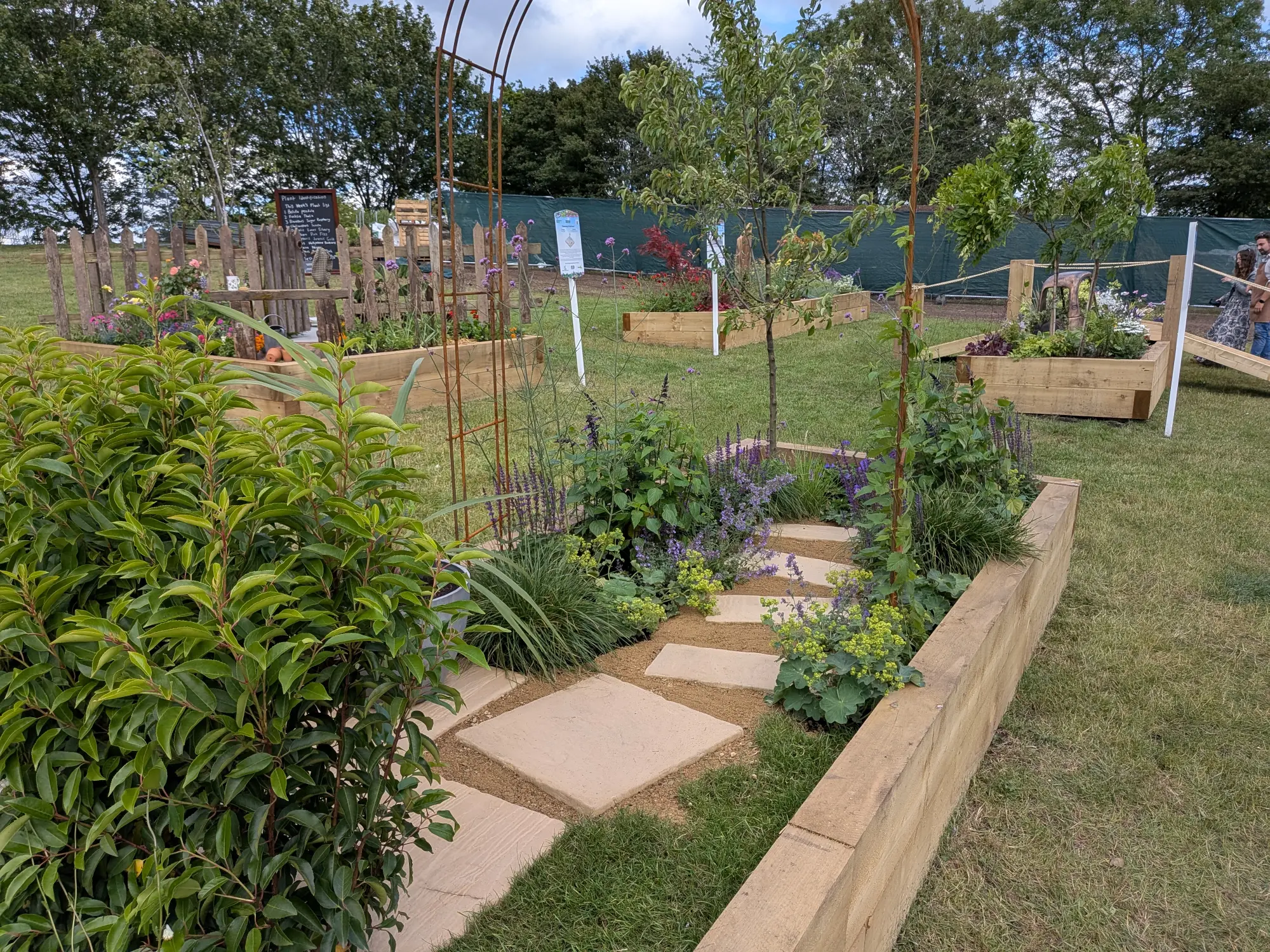 A beautiful border garden features a winding stepping stone path, naturalistic green and purple planting, timber raised beds, and rusty metal arch trellises.