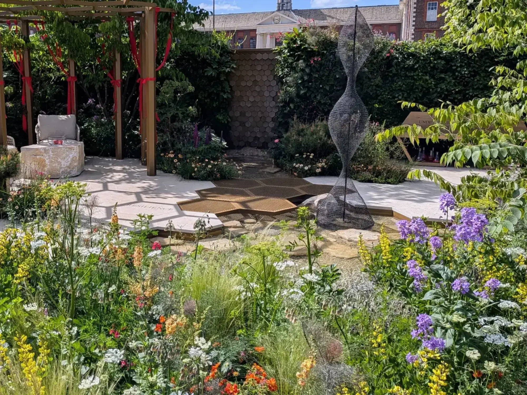 A show garden features a wire DNA helix sculpture, hexagonal bronze paving with a water feature, a wooden pergola draped with red ribbons, and abundant, colourful wildflowers.