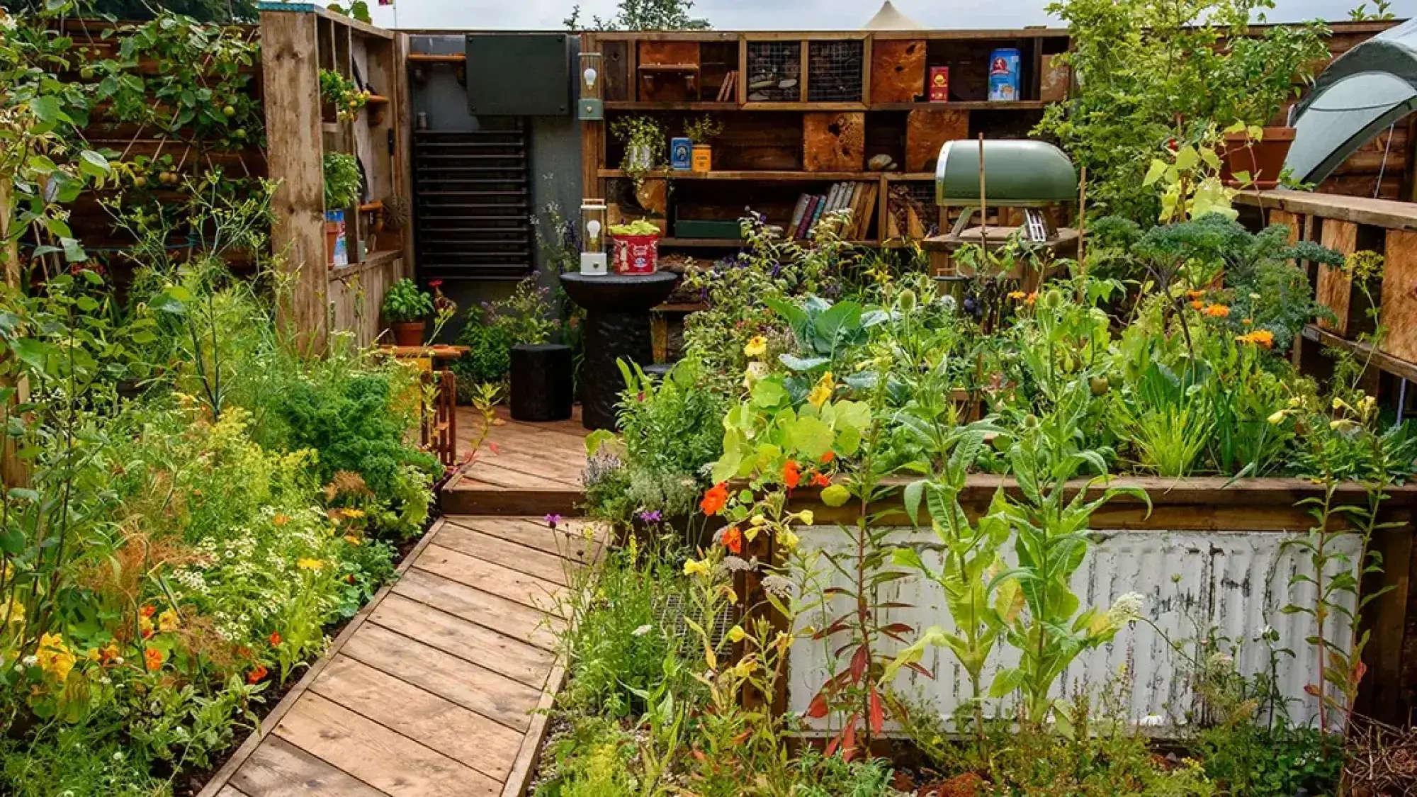 A rustic terrace garden features a timber decking path, raised beds with edible plants and orange flowers, and a reclaimed wooden shelving unit with a pizza oven.