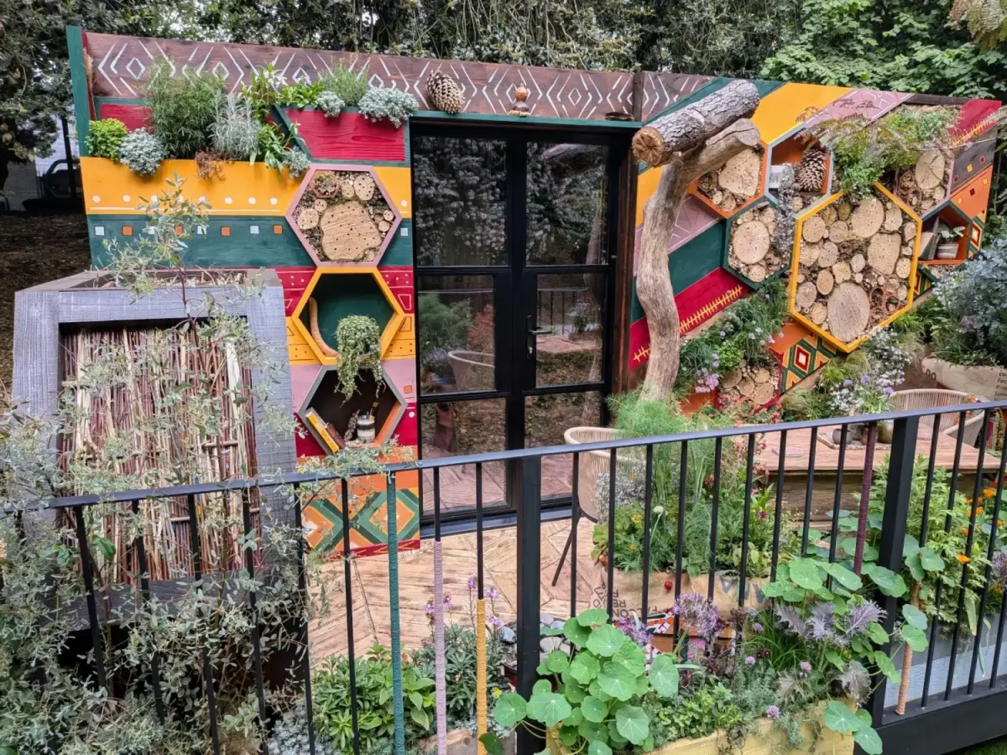 A vibrant balcony garden features a colourful geometric wall with hexagonal bee habitats of wood slices, a central door, and lush pollinator plants.