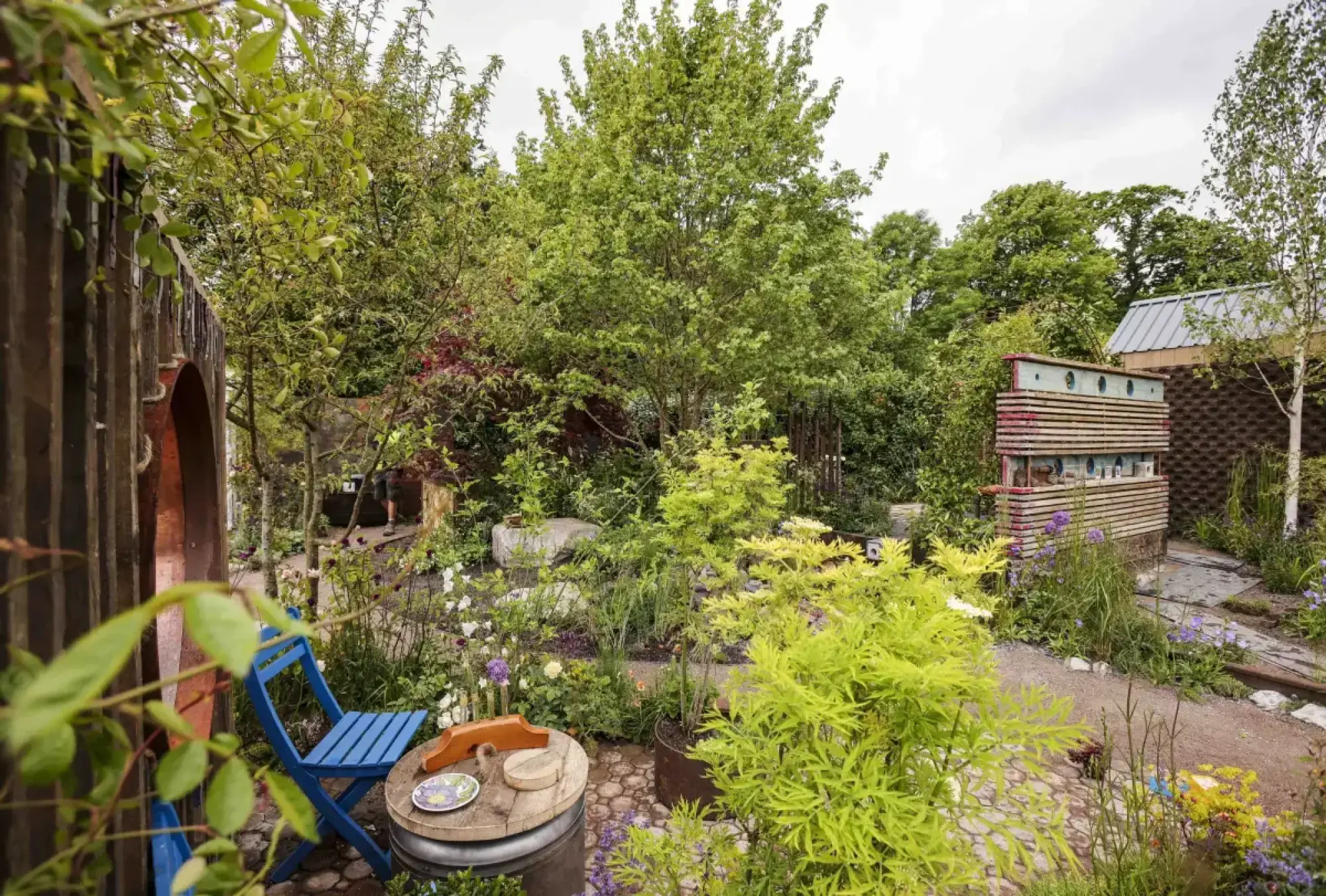 A naturalistic medium show garden with a stacked wooden bug hotel, a blue chair, barrel table, and vibrant lime green foliage among mixed herbaceous plants.