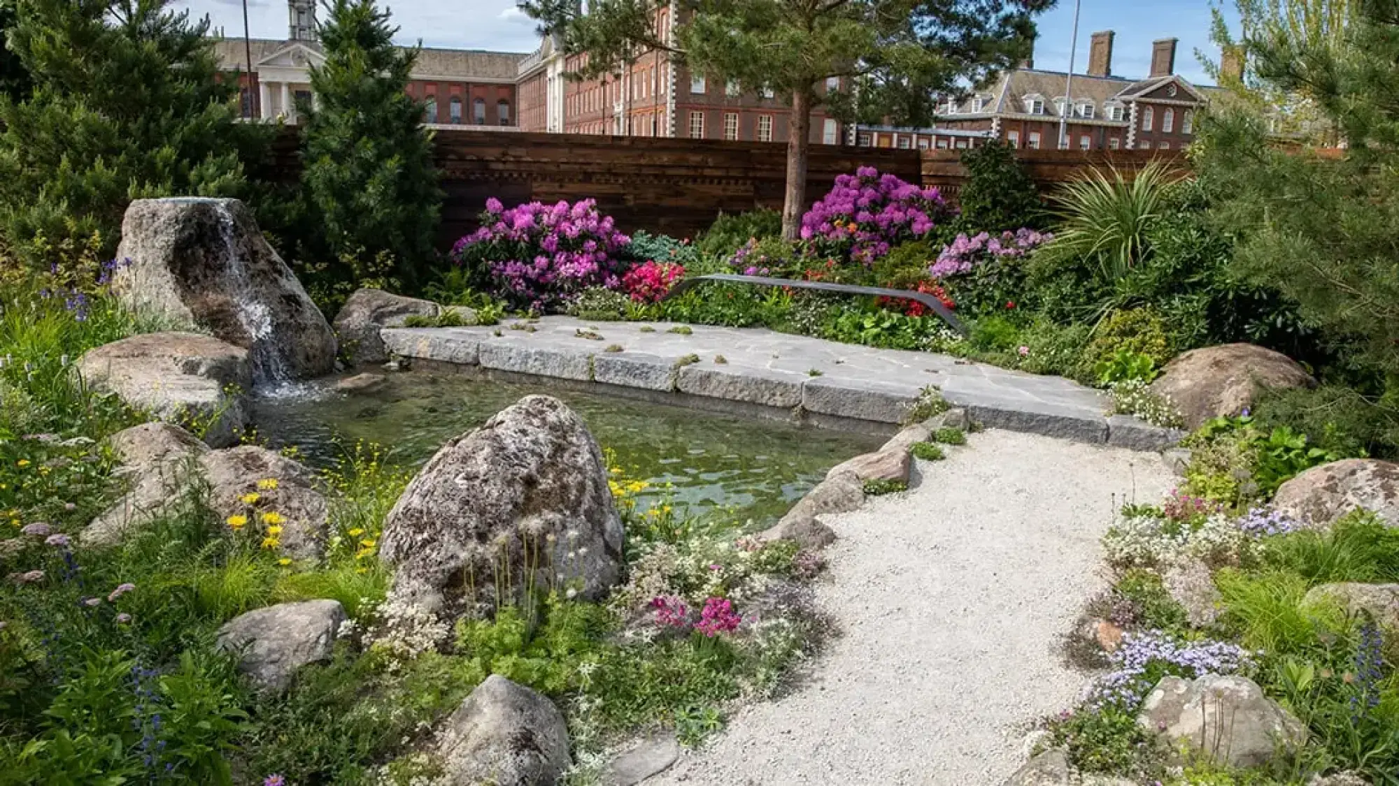 A tranquil sanctuary garden features a rock waterfall into a granite pond, bordered by a gravel path and alpine planting with pink rhododendrons.