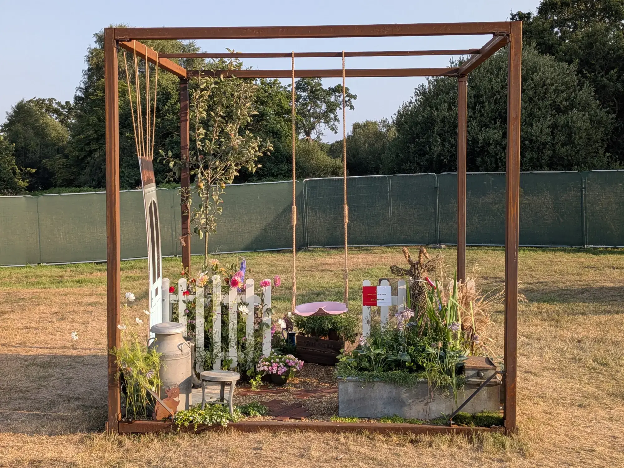 A rustic country garden set within a rusted metal cube, featuring a pink swing, a white picket fence, an apple tree, mixed flowers, and a brick path.