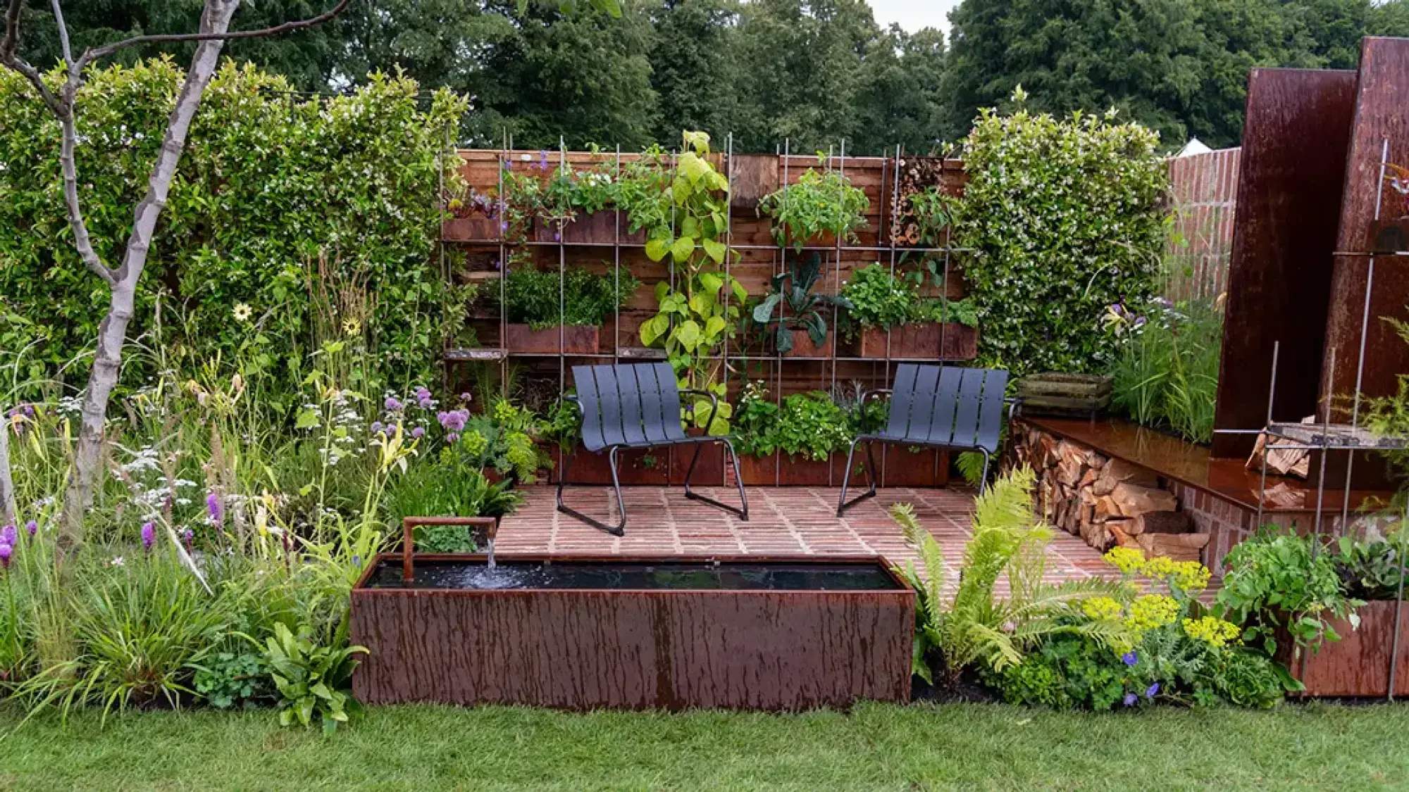 A contemporary slim garden with a corten steel water feature, a brick patio, dark chairs, and a vertical wall of plants in rusty metal containers.