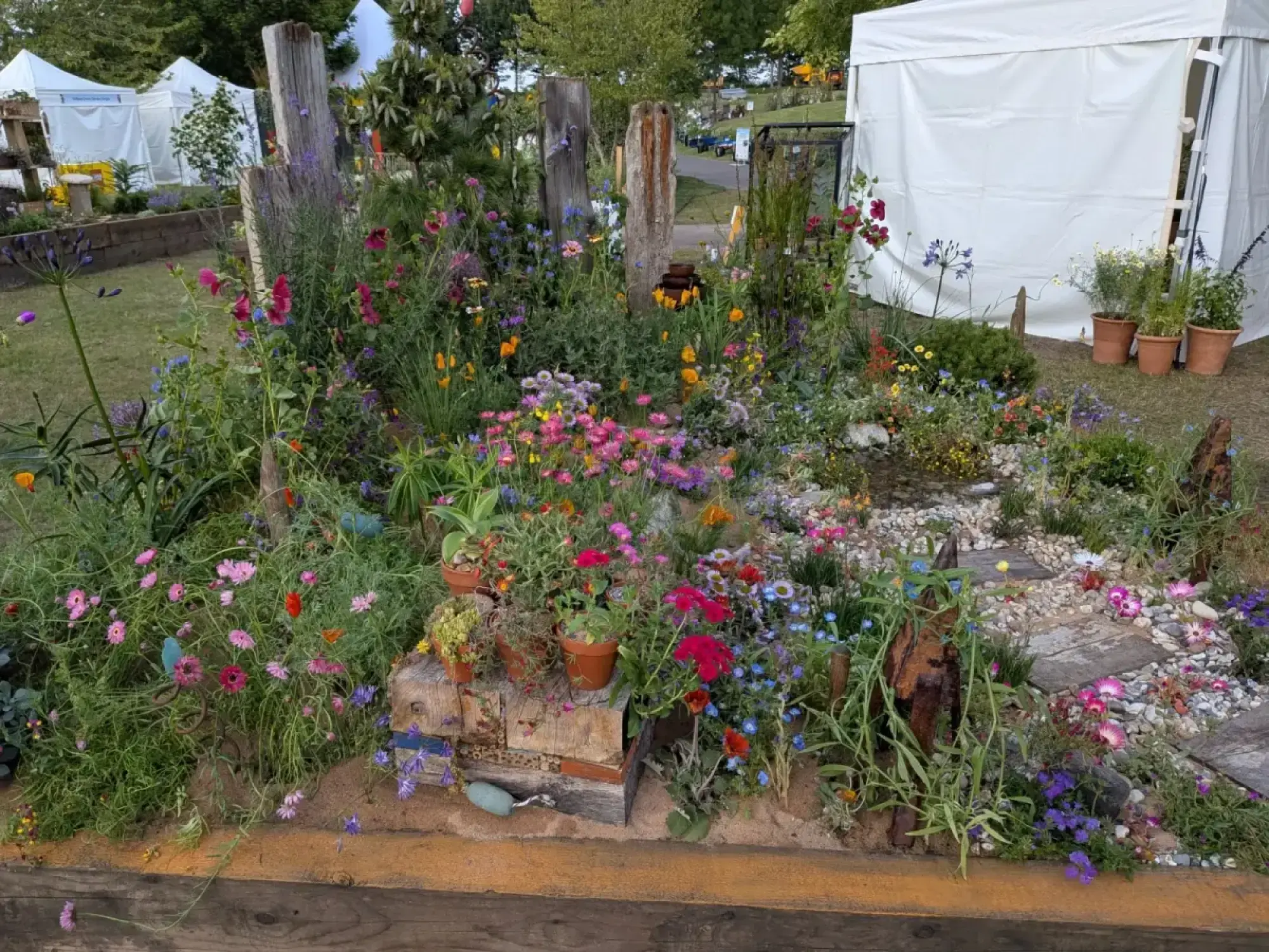 A vibrant dry border garden densely planted with pink, purple, blue, yellow, and orange flowers, featuring weathered timber posts and a pebble path.