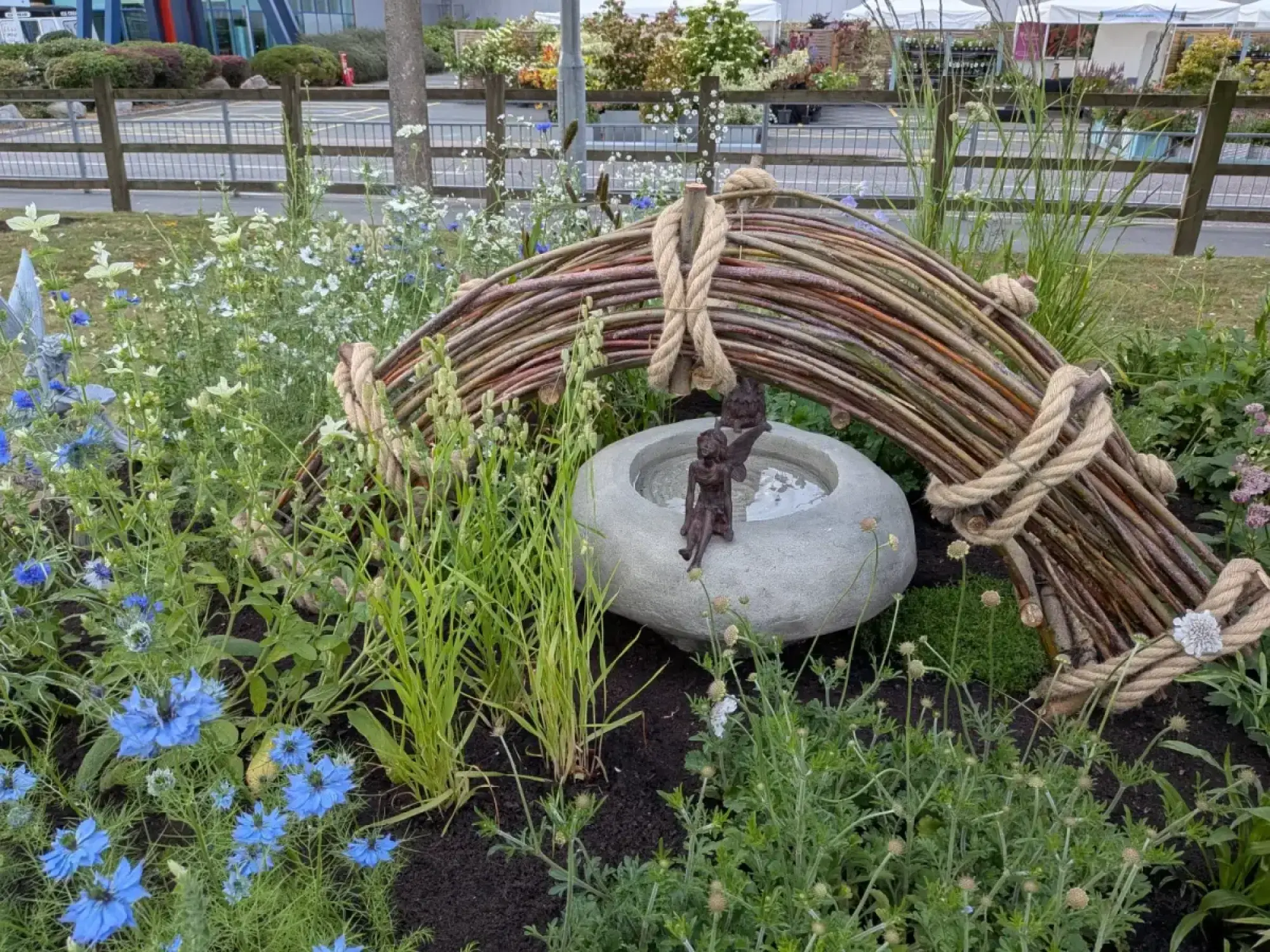 A beautiful border features a woven willow arch tied with rope, sheltering a stone bird bath with bronze child figures, amidst blue Nigella and white wildflowers.
