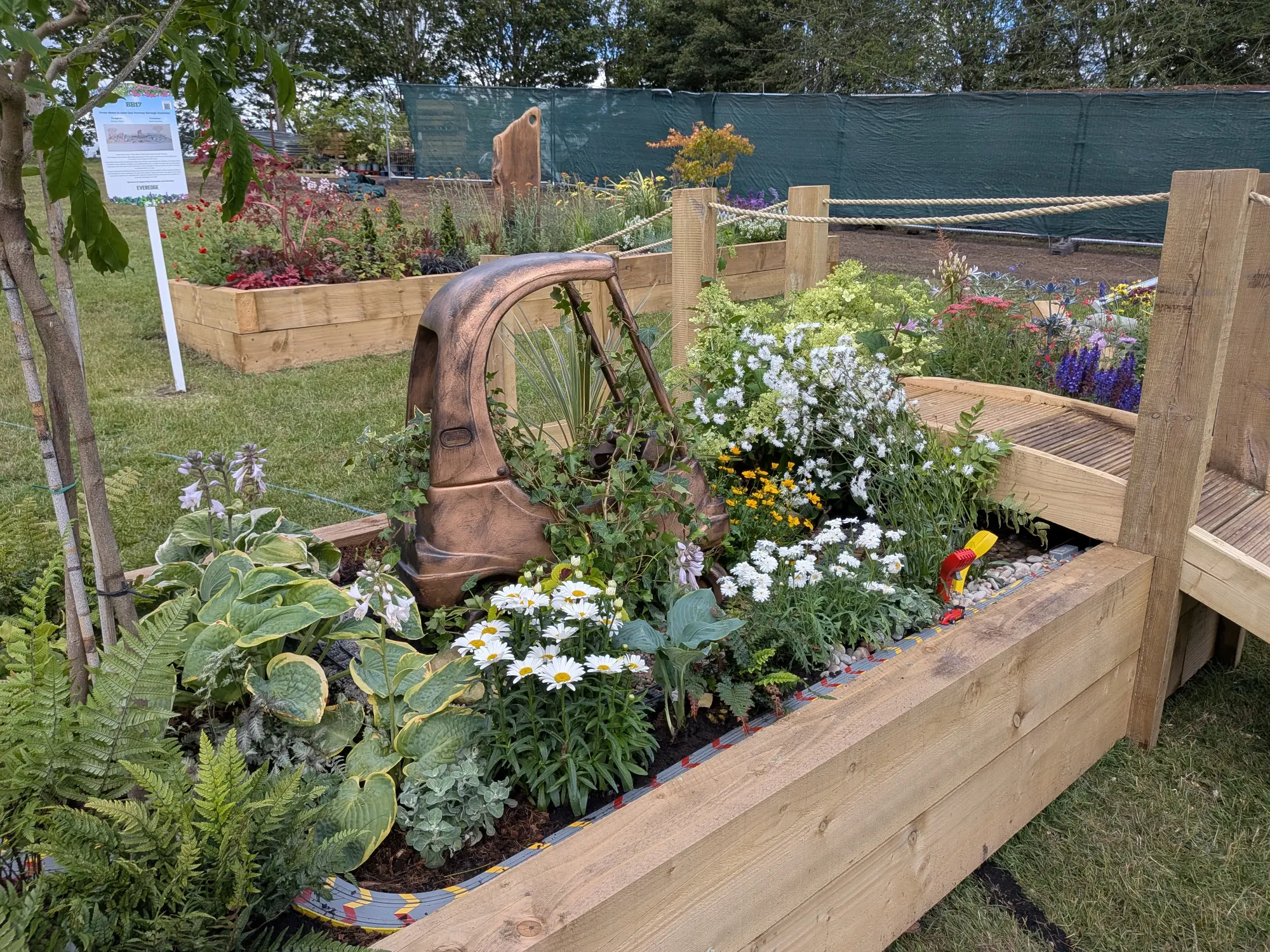 A playful raised timber border garden features a copper car sculpture, a toy car track, white and yellow flowers, ferns, and hostas.